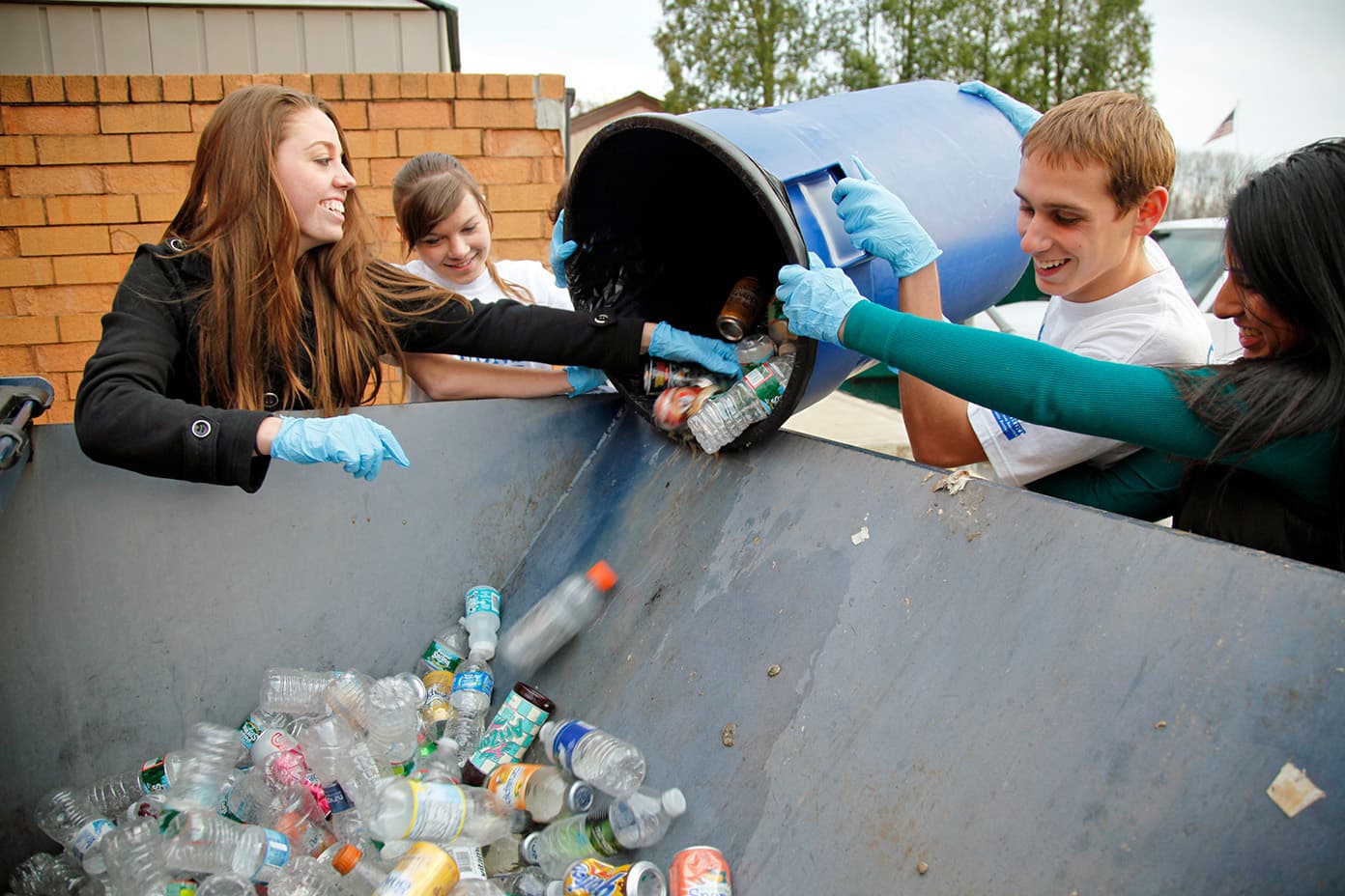 Four teenagers in blue gloves tip a blue wheelie bin of bottles and cans over the lip of a large metal skip; plastic bottles tumble down onto the pile already inside.