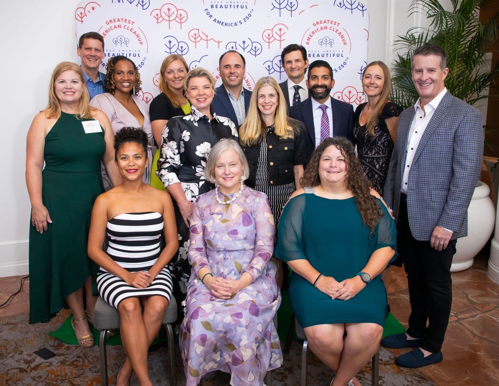 Thirteen people in evening dress pose in three rows, three of them seated on chairs, in front of a white step-and-repeat printed with red and blue tree motifs and the GREATEST AMERICAN CLEANUP seal.