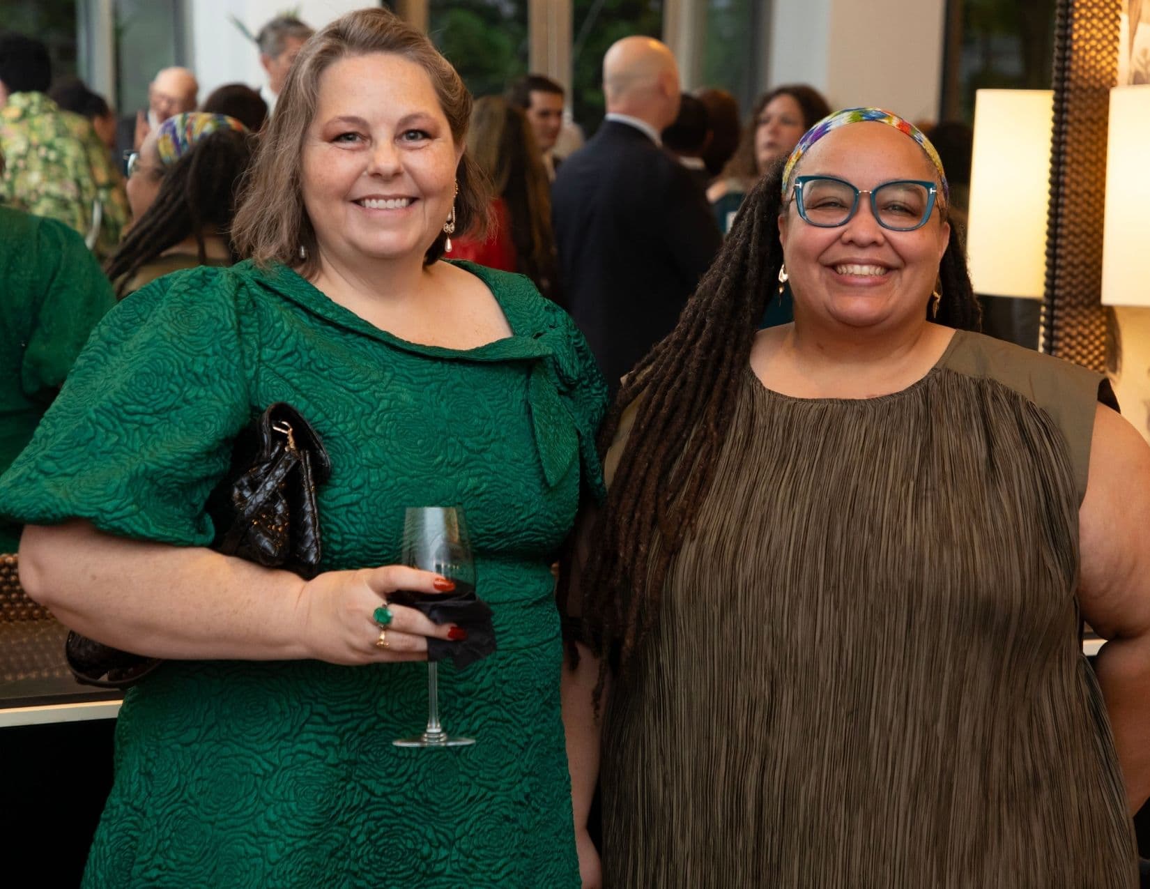Two women pose at a crowded reception: one in a textured emerald green dress holding a glass of red wine and a quilted clutch, the other in a pleated olive shift with long locs, a patterned headband and blue glasses.