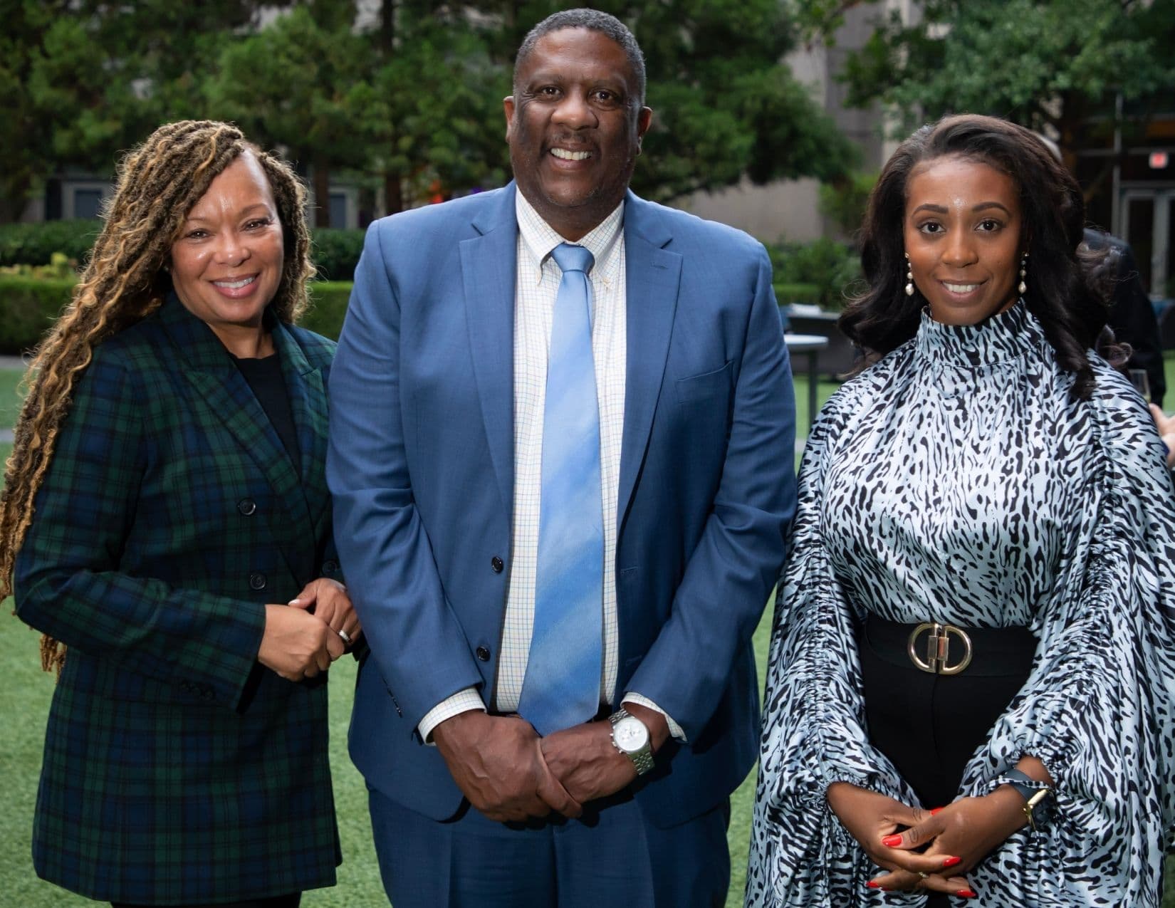 Three people stand arm in arm on a lawn at dusk: a woman in a green and navy tartan blazer, a man in a mid-blue suit with a pale blue tie, and a woman in a black and white animal-print top with a gold belt buckle.