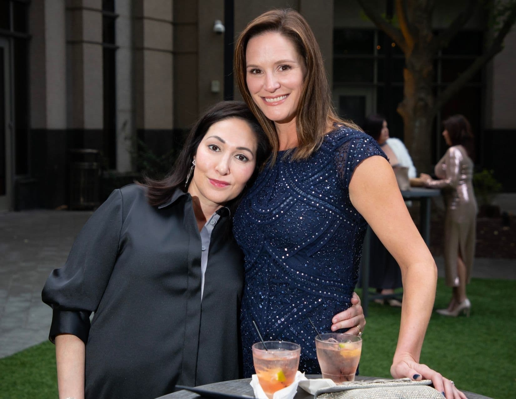 Two women lean together at an outdoor drinks table on artificial grass, one in a black satin shirt dress, the other in a navy sequinned dress; two cocktails and a beaded clutch sit on the table in front of them.