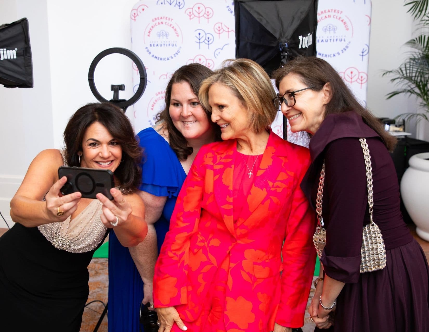 Four women crowd together for a phone selfie in front of a branded backdrop and ring light. The woman at left holds the phone out; the centre figure wears a bright pink and orange floral jacket over a matching dress.