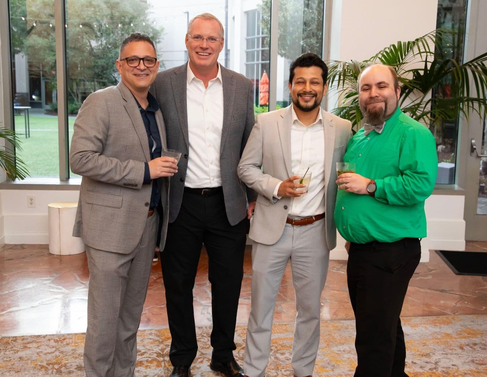 Four men stand shoulder to shoulder in a marble-floored atrium holding drinks: two in checked grey jackets, one in a pale beige blazer, and one in a bright green shirt with a grey bow tie.