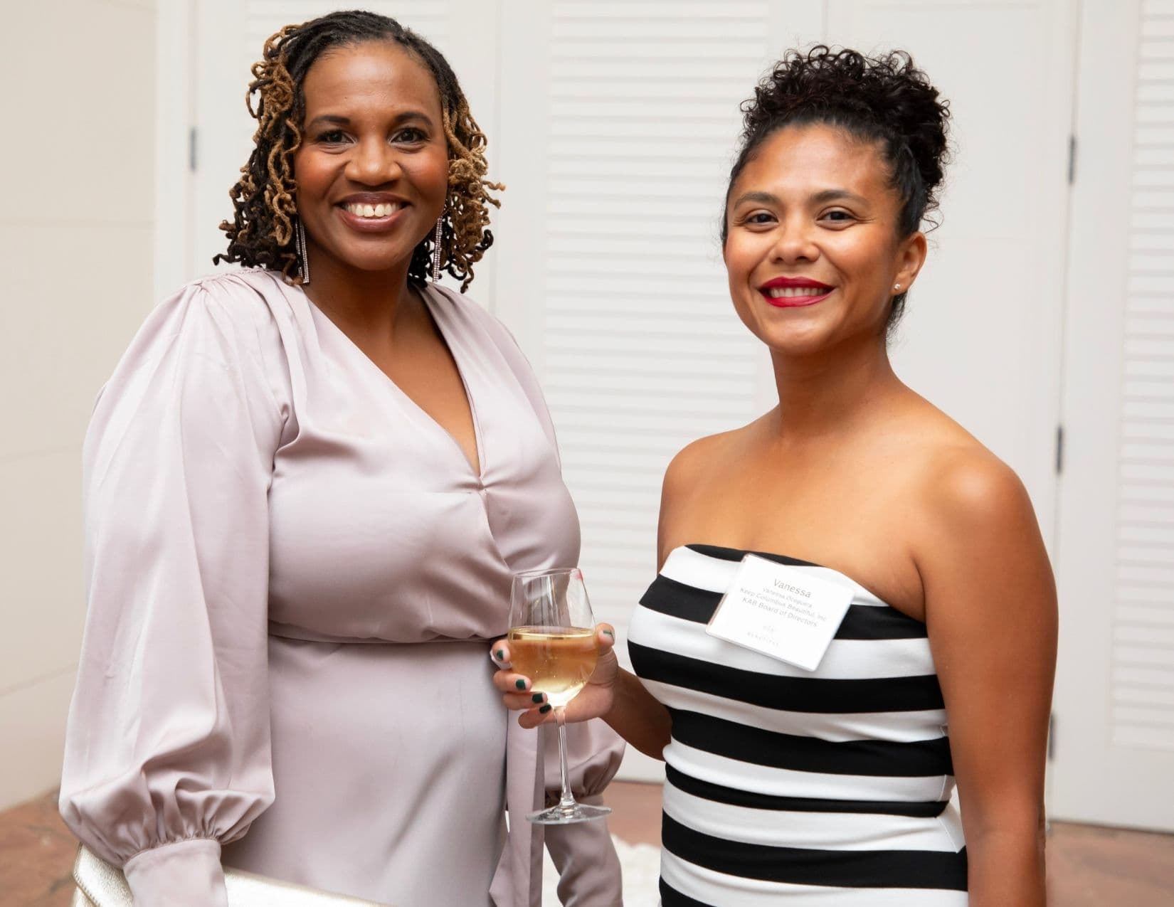 Two women pose beside louvred doors. The left wears a blush satin blouse and holds a glass of white wine; the right wears a black and white horizontally striped strapless dress with a name badge reading Vanessa, Keep Columbus Beautiful Inc., KAB Board of Directors.