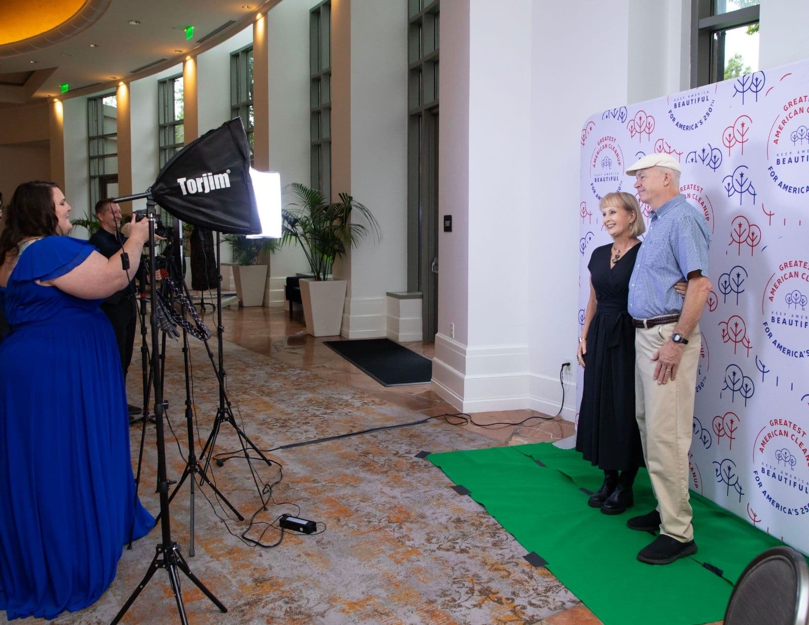 A wide hotel lobby view: a woman in a cobalt blue gown and a photographer work a Torjim softbox and tripod at the left while an older couple pose on a green mat against a patterned step-and-repeat at the right.