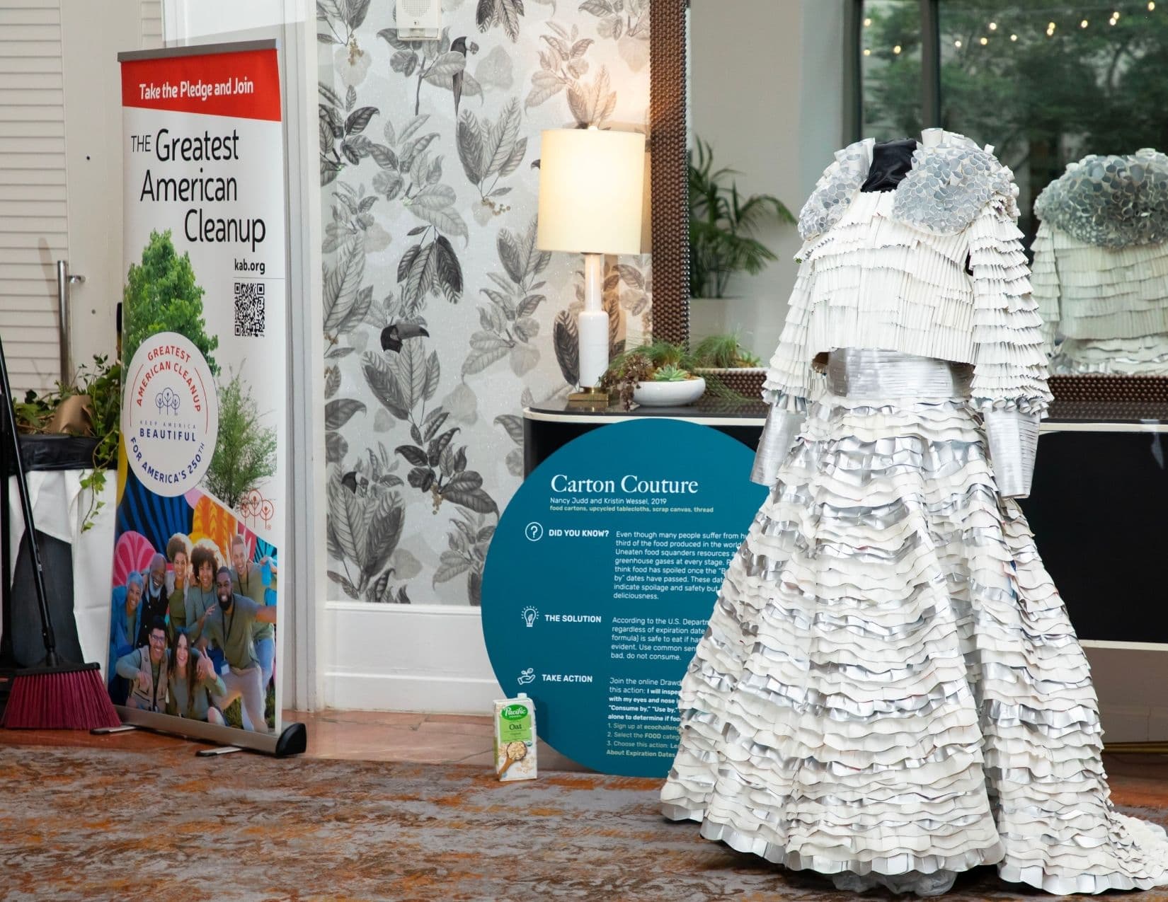 A ruffled ballgown built from folded white paper stands on a mannequin beside a teal circular sign headed Carton Couture, Nancy Judd and Kristin Wessel, 2019, next to a roll-up banner reading Take the Pledge and Join THE Greatest American Cleanup.