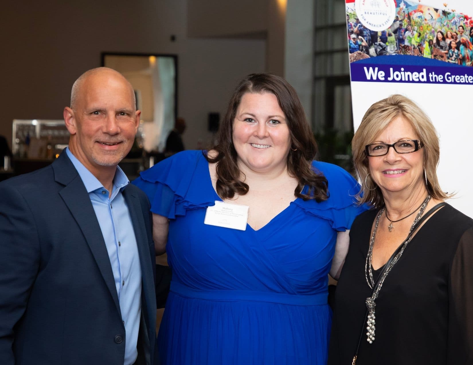 Three people pose in front of a banner reading We Joined the Greatest: a bald man in a navy jacket, a woman in a royal blue gown wearing a badge printed Mallory, and a woman in black with a long tasselled necklace.