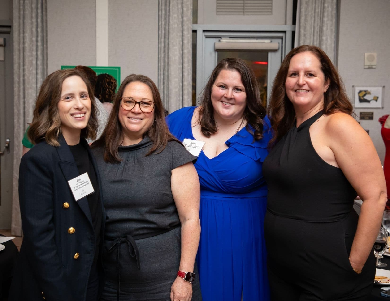 Four women stand together in a banqueting room, two wearing name badges reading Erin and Mallory. From left: a navy blazer with gold buttons, a grey shift dress, a royal blue gown, and a black halter jumpsuit.