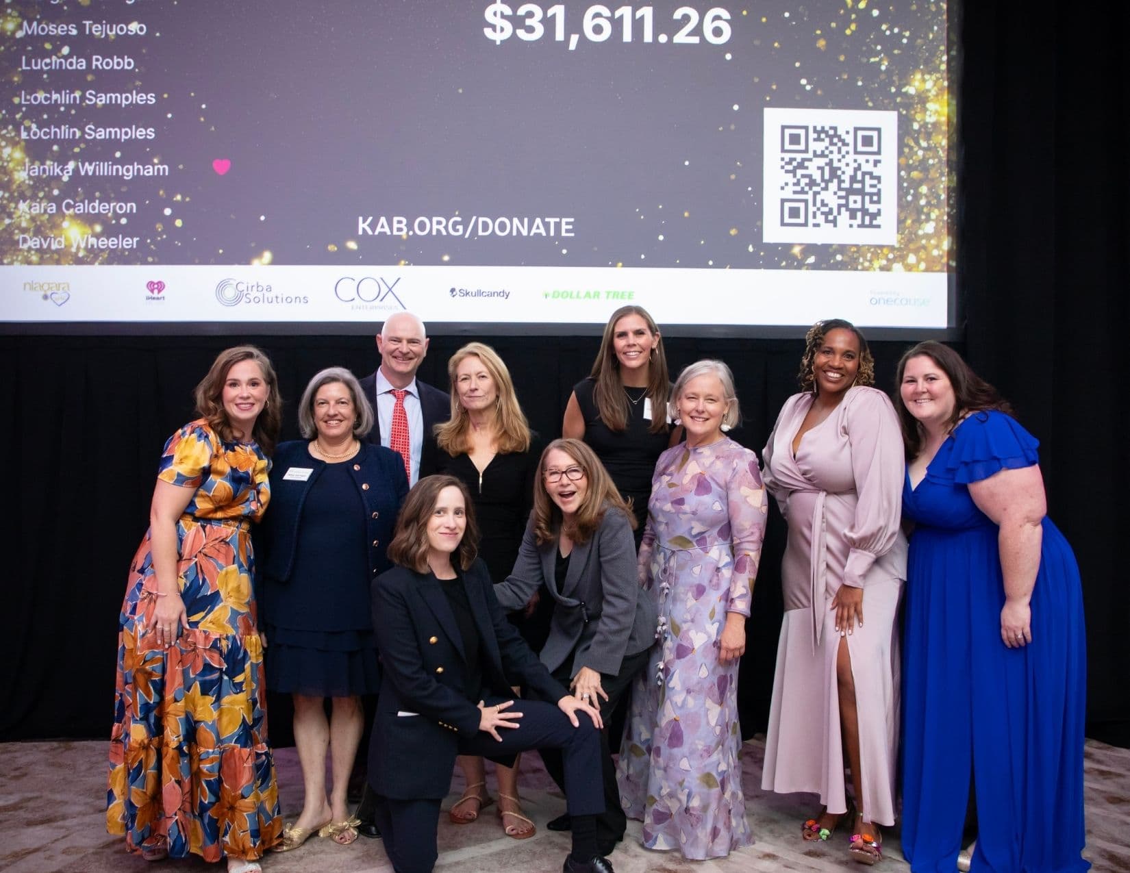 Eleven people pose on a low stage below a projection screen showing a donor list, the total $31,611.26, a QR code, KAB.ORG/DONATE and sponsor logos including Niagara, iHeart, Cirba Solutions, Cox Enterprises, Skullcandy and Dollar Tree.