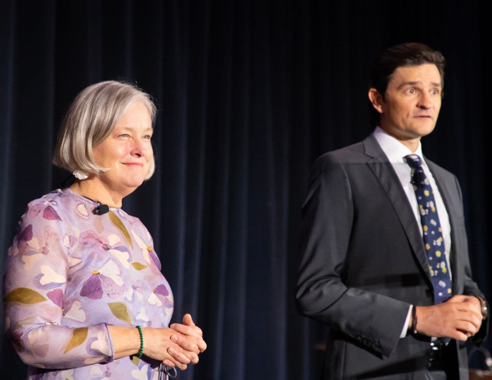 A woman with a silver-grey bob in a lilac floral dress stands smiling at the left of a dark stage curtain, hands clasped, while a man in a charcoal suit and navy floral tie stands apart from her at the right.