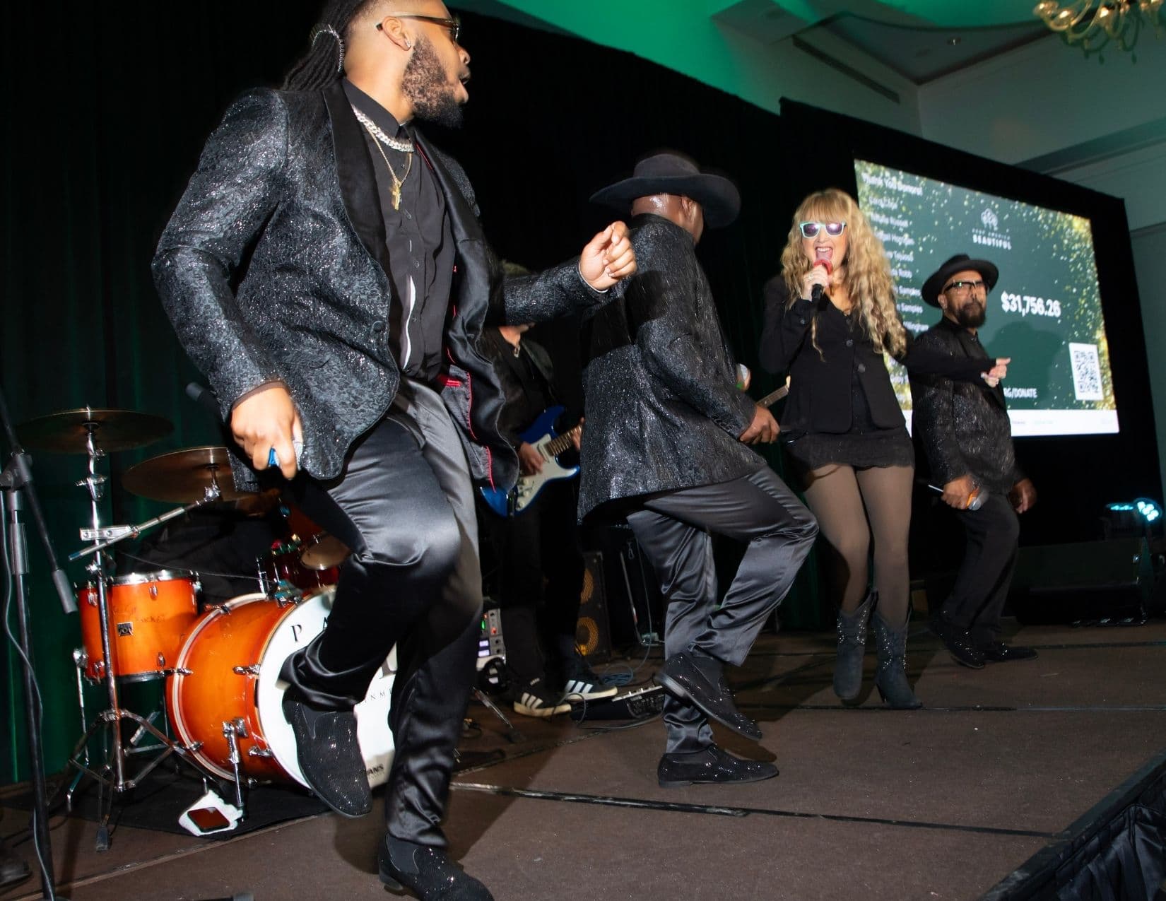 A band performs on a small stage: two men in glittering black jackets dance mid-step, a woman with curly blonde hair and white sunglasses sings into a microphone, and a drum kit and blue bass guitar sit behind them.