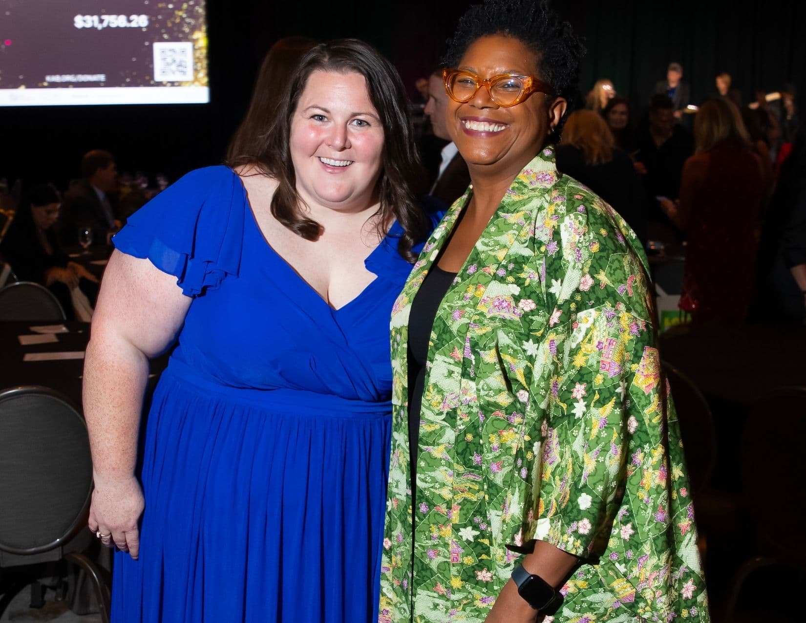 Two women stand together at a banquet: one in a royal blue chiffon gown with flutter sleeves, the other in orange-framed glasses and a long green floral duster over a black top.