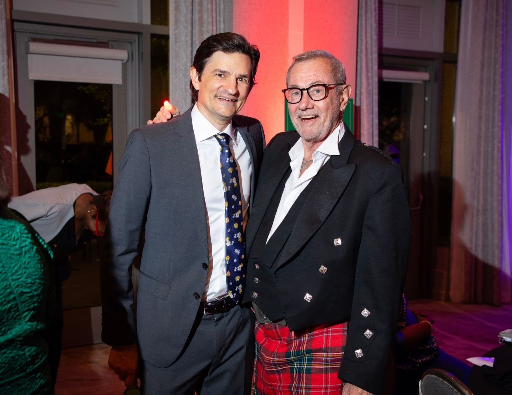 Two men pose in a room lit red and purple: one in a grey suit with a navy floral tie, the other older, in black-framed glasses, an open white shirt and a black jacket with silver buttons over a red tartan kilt.