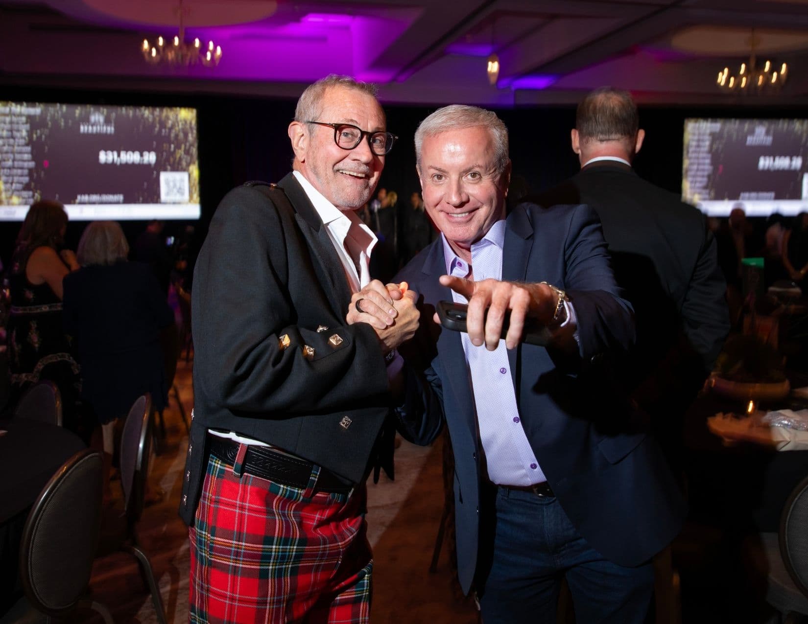 In a ballroom, a man in a red tartan kilt and buttoned black jacket clasps hands with a white-haired man in a navy blazer and lilac shirt who holds out a phone; screens behind them show a fundraising total.