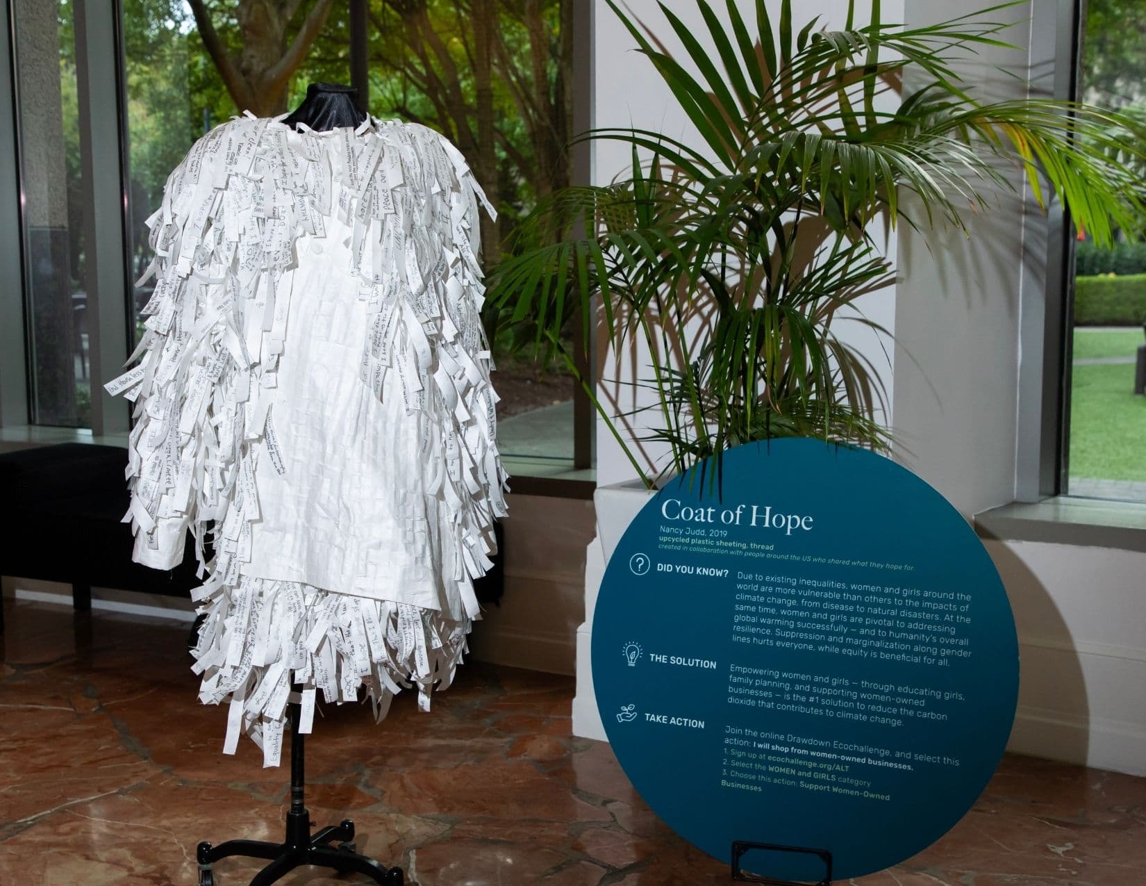 A pale coat covered in hundreds of small handwritten paper tags hangs on a dress form beside a potted palm and a teal circular sign headed Coat of Hope, Nancy Judd, 2019, upcycled plastic sheeting, thread.