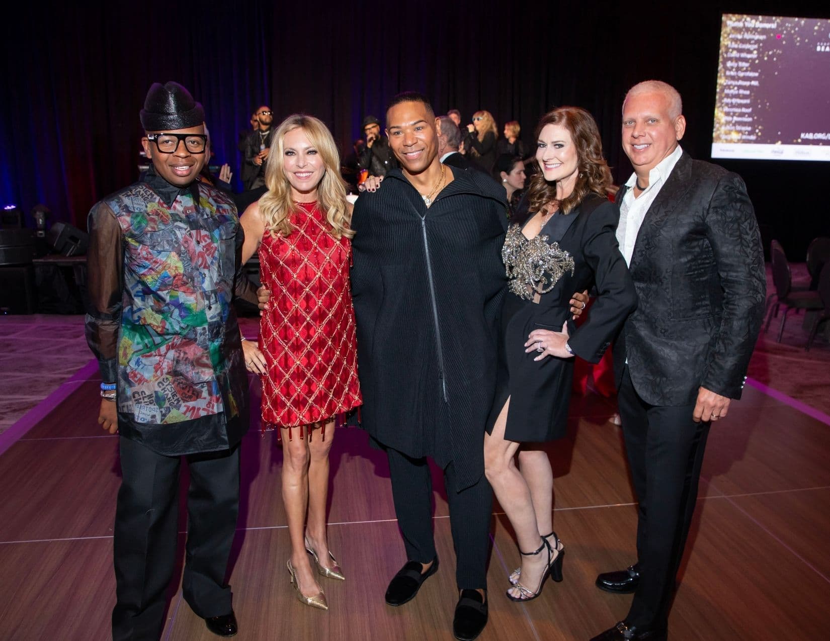 Five people pose on a wooden dance floor: a man in a printed plastic-patchwork jacket and black hat, a woman in a red and gold beaded dress, a man in a black pleated cape-style zip top, a woman in a black jacket dress with a jewelled bodice, and a man in a black brocade jacket.