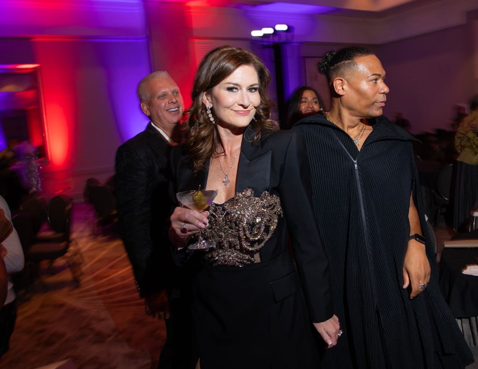 A woman in a black jacket dress with an ornate crystal bodice walks through a ballroom holding a martini garnished with an olive, alongside a man in a black pleated cape top; another man in a brocade jacket laughs behind her.
