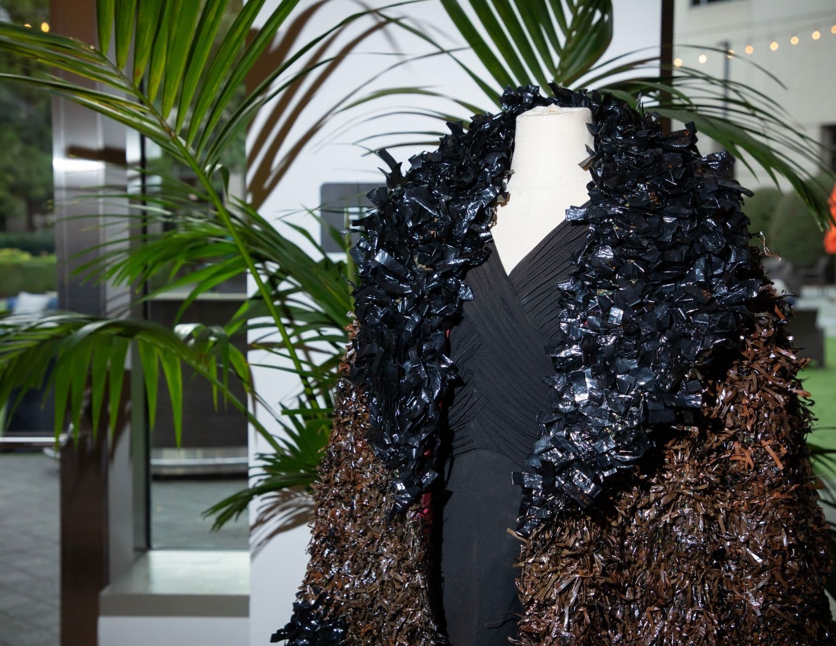 Close view of a mannequin wearing a black draped bodice under a shaggy coat made from shredded black plastic bags at the shoulders and shredded bronze and brown plastic below, with a potted palm in the foreground.
