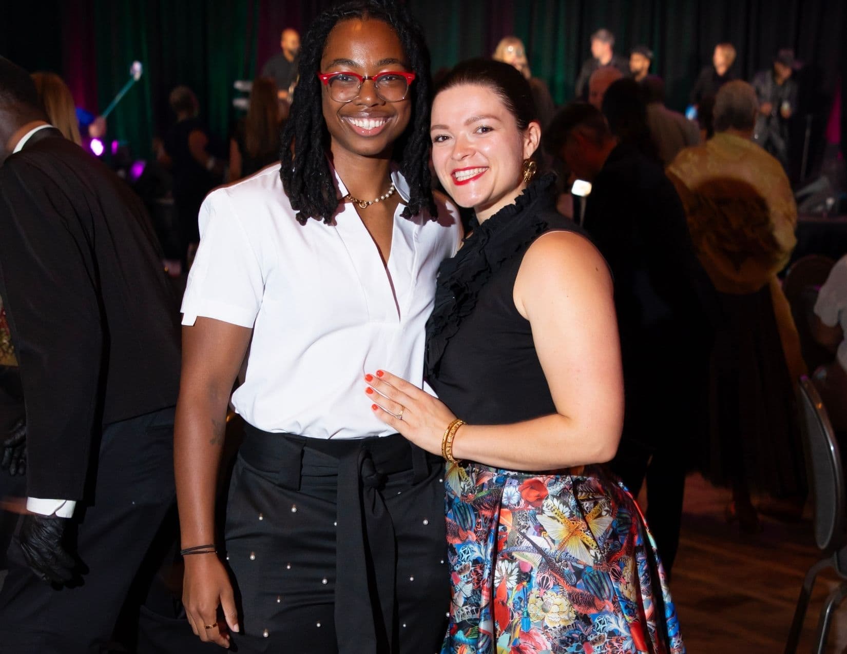 Two women pose at a busy evening reception: one with shoulder-length locs, red-framed glasses and a white short-sleeved shirt with black trousers, the other in a black sleeveless ruffled top and a brightly printed pleated skirt.