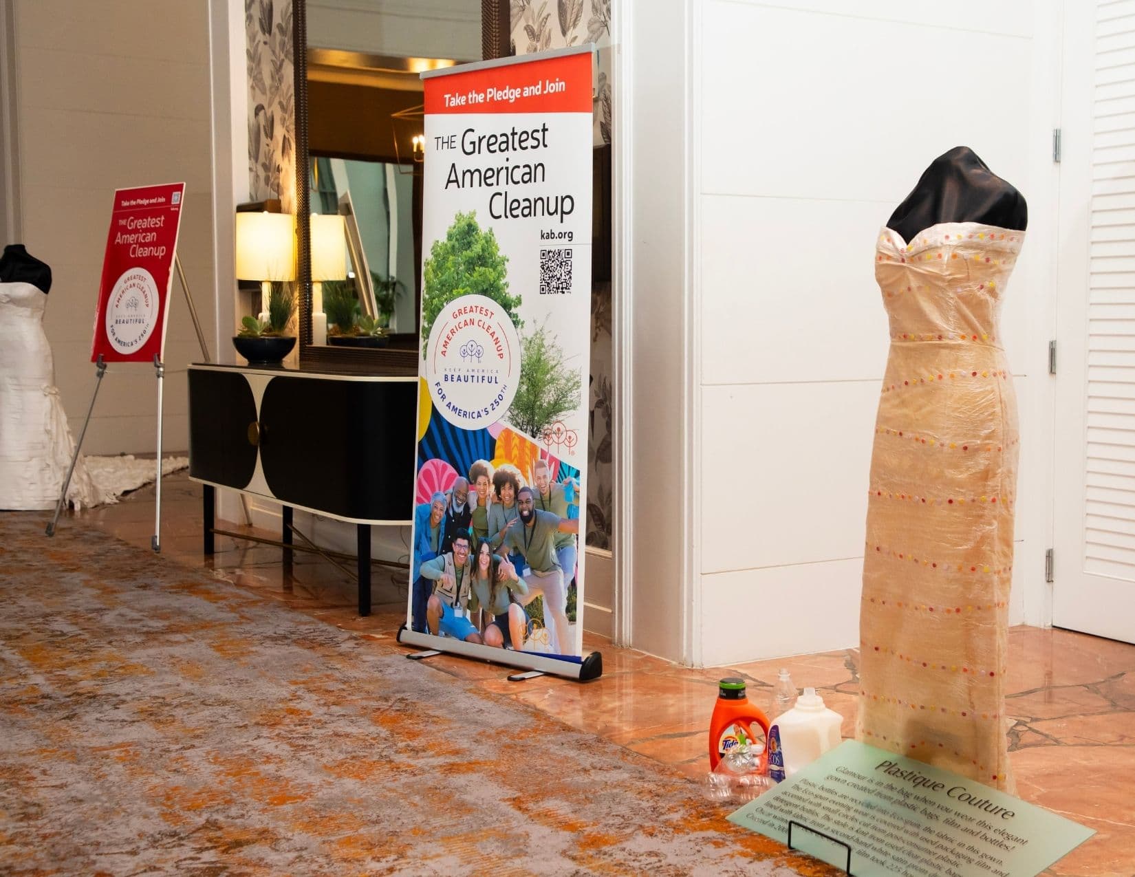 A hotel corridor display: a floor-length gown made from translucent beige plastic sheeting studded with coloured dots stands beside a card reading Plastique Couture, with detergent bottles at its base and a Greatest American Cleanup roll-up banner nearby.