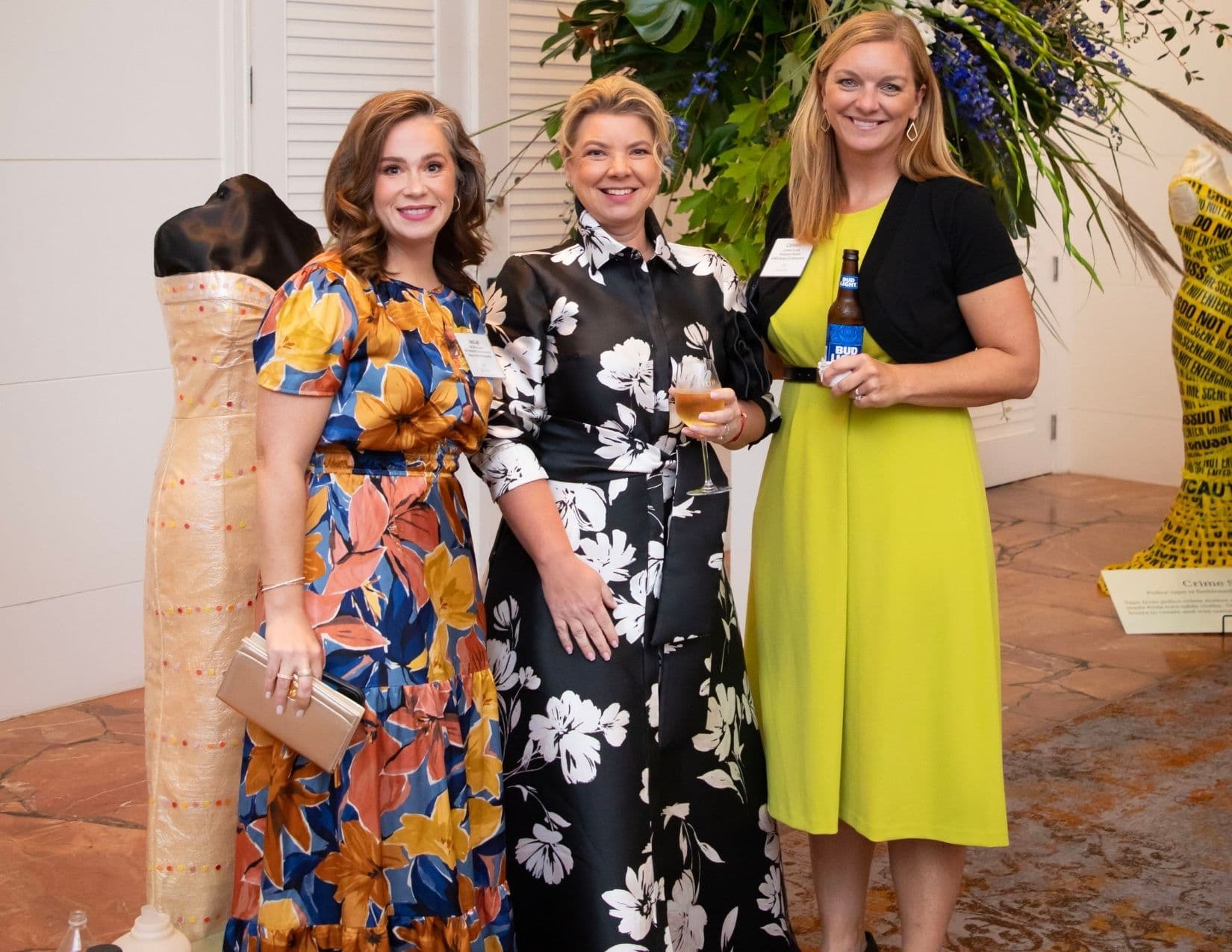Three women pose between two mannequins, one dressed in beige plastic sheeting and one in yellow caution tape. They wear a blue and orange floral dress, a black and white floral gown, and a lime green dress with a bottle of Bud Light.