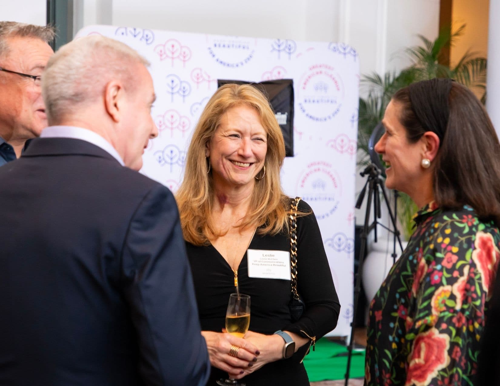 A woman with long strawberry-blonde hair laughs mid-conversation between two other guests, wearing a black zip-front dress and a chain-strap bag, with a name badge reading Leslie McClain, VP of Communications, Keep America Beautiful.