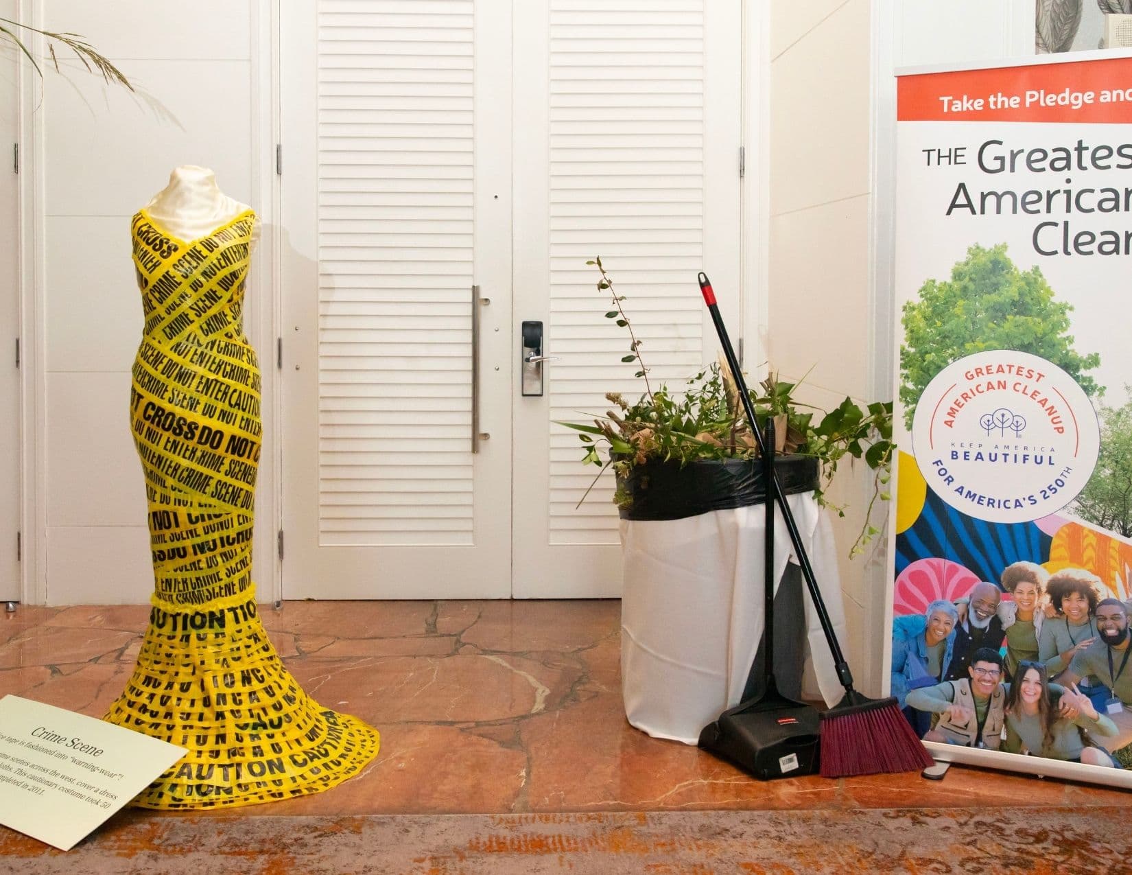 A mermaid-cut gown wrapped entirely in yellow CAUTION and CRIME SCENE DO NOT CROSS tape stands on a mannequin beside a card titled Crime Scene, with a bin, broom and dustpan and a Greatest American Cleanup banner to the right.