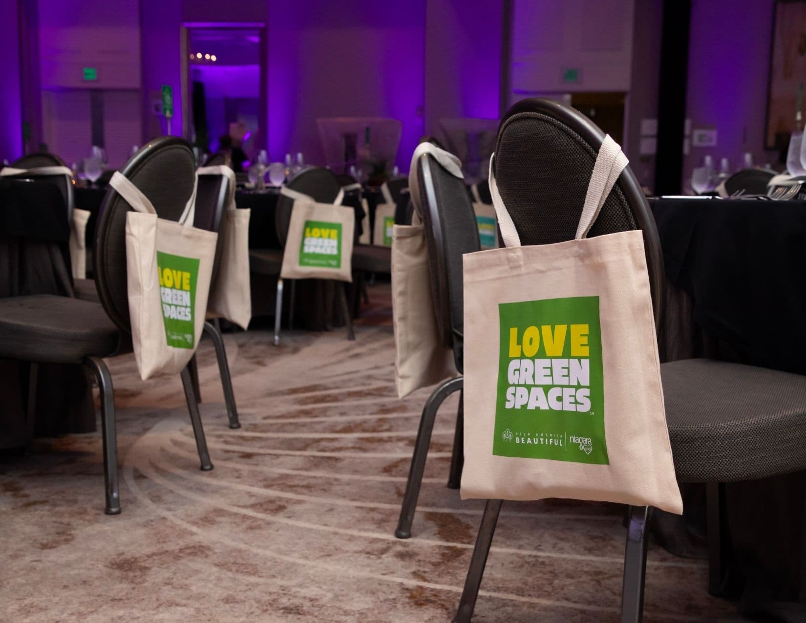 Rows of banquet chairs in a purple-lit ballroom, each hung with a natural canvas tote bag printed LOVE GREEN SPACES in yellow and white on a green panel above the Keep America Beautiful and Niagara Cares logos.