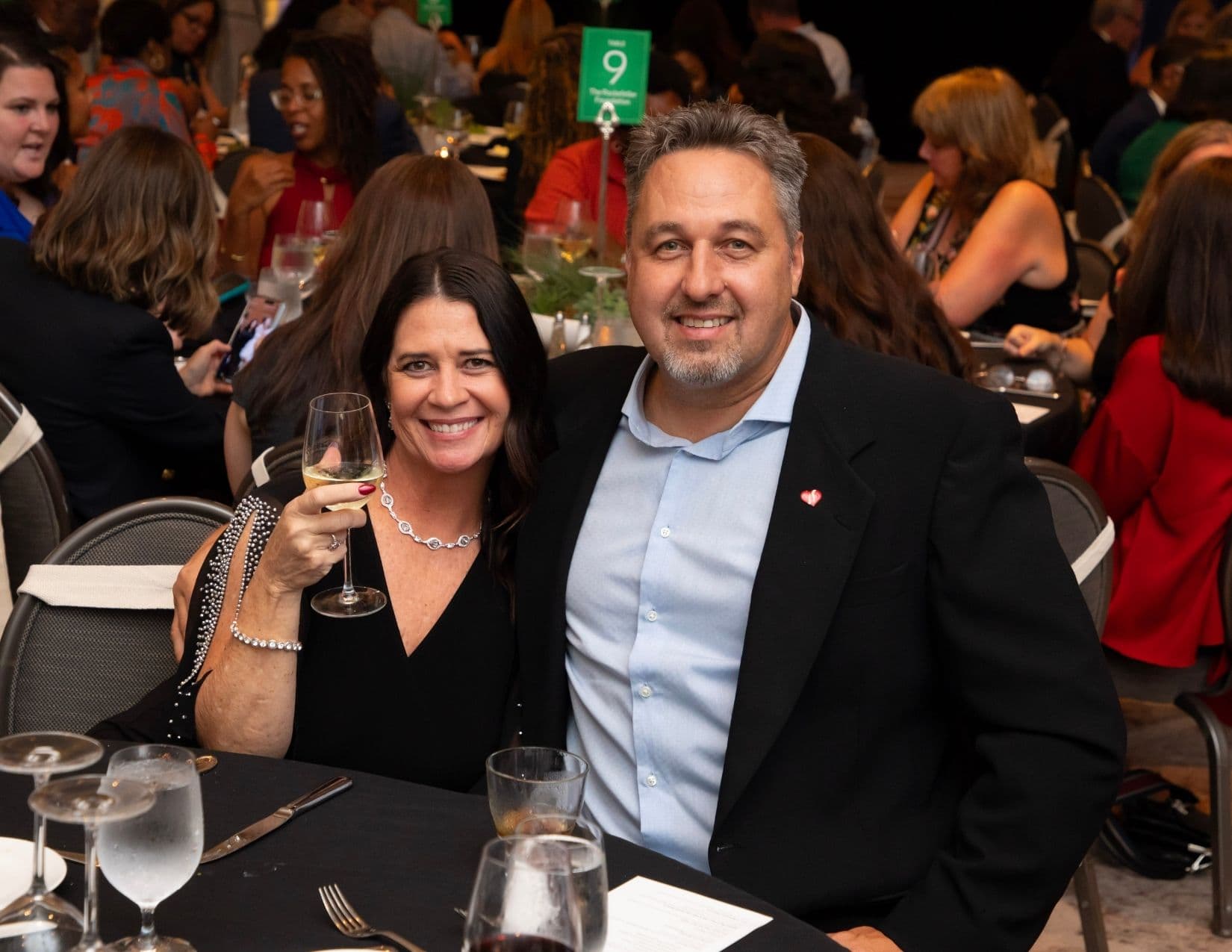 A couple pose at a banquet table beneath a green table marker numbered 9; the woman in black raises a glass of white wine, the man beside her wears a black jacket over a pale blue shirt with a small red pin.