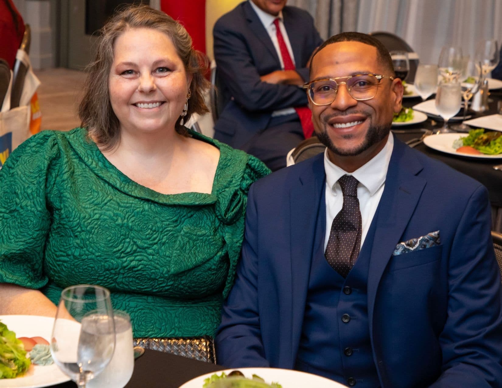 A woman in a textured emerald green dress and a man in a navy three-piece suit with a patterned tie and pocket square sit side by side at a banquet table, salad plates and wine glasses in front of them.