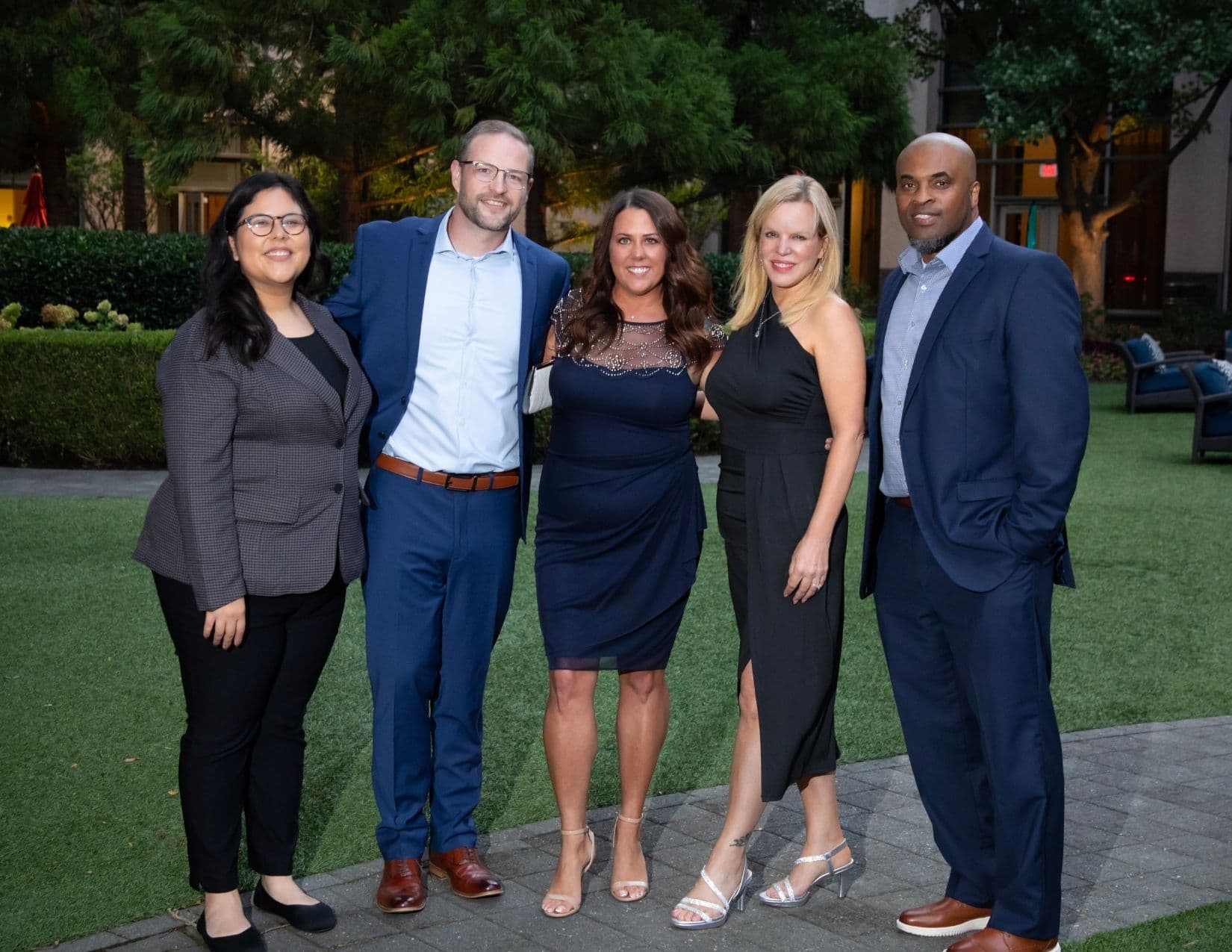 Five people stand in a line on a lawn in the evening: a woman in a checked grey blazer, a man in a blue suit, a woman in a navy embellished dress, a woman in a black halter dress with a thigh split, and a man in a navy suit.