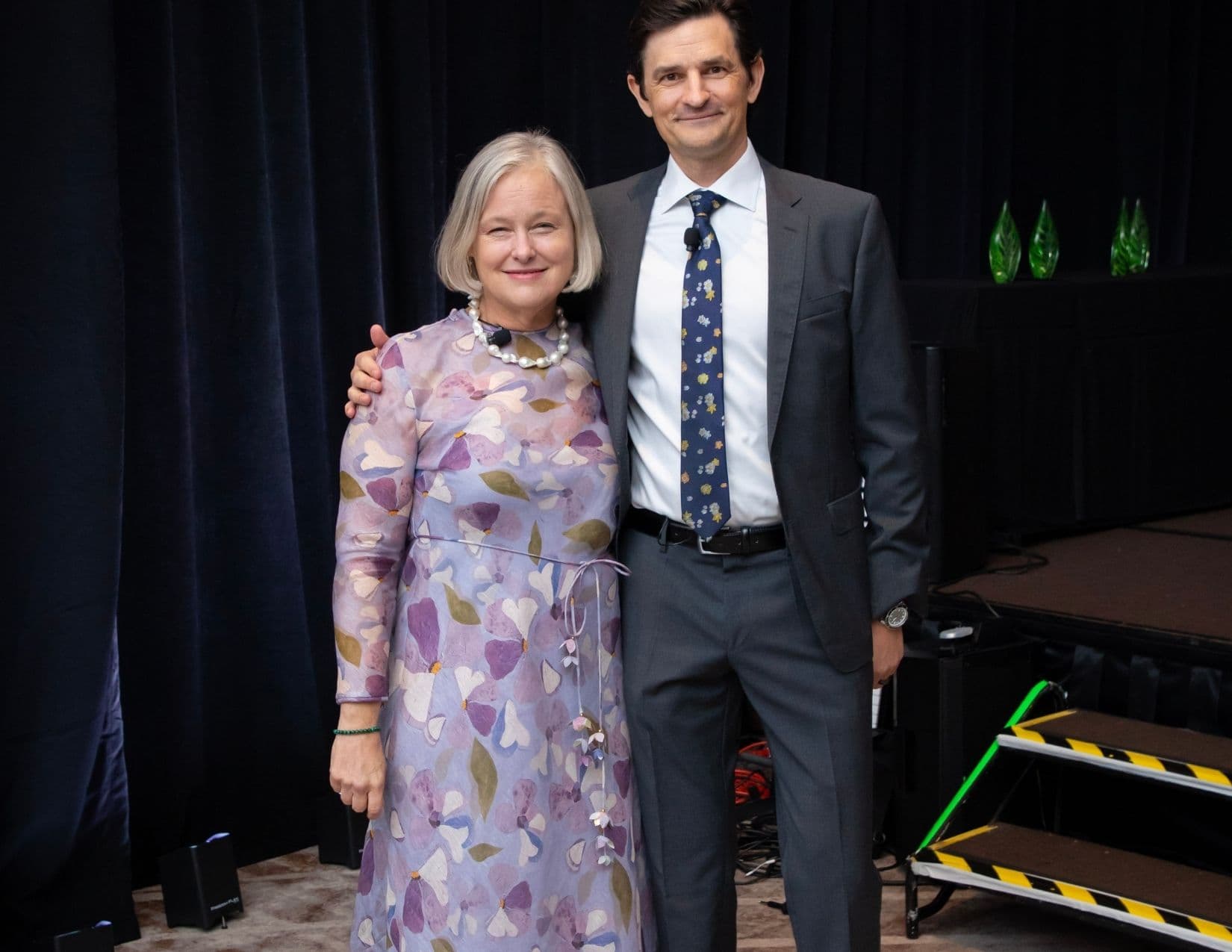 A woman in a lilac floral dress with a pearl necklace stands close beside a taller man in a charcoal suit and navy floral tie who has his arm around her shoulders; green glass awards line a table behind them.