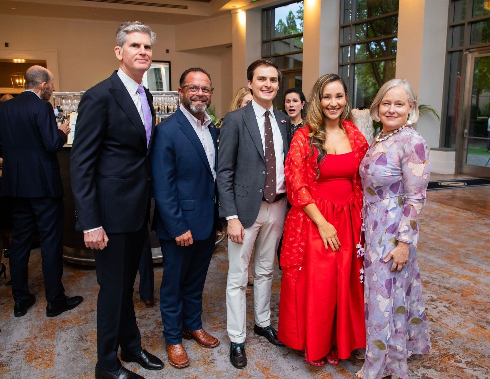 Six people pose near a hotel bar: three men in suits, a young man in a grey blazer and cream trousers, a woman in a red dress with a red printed wrap, and a woman in a lilac floral gown.