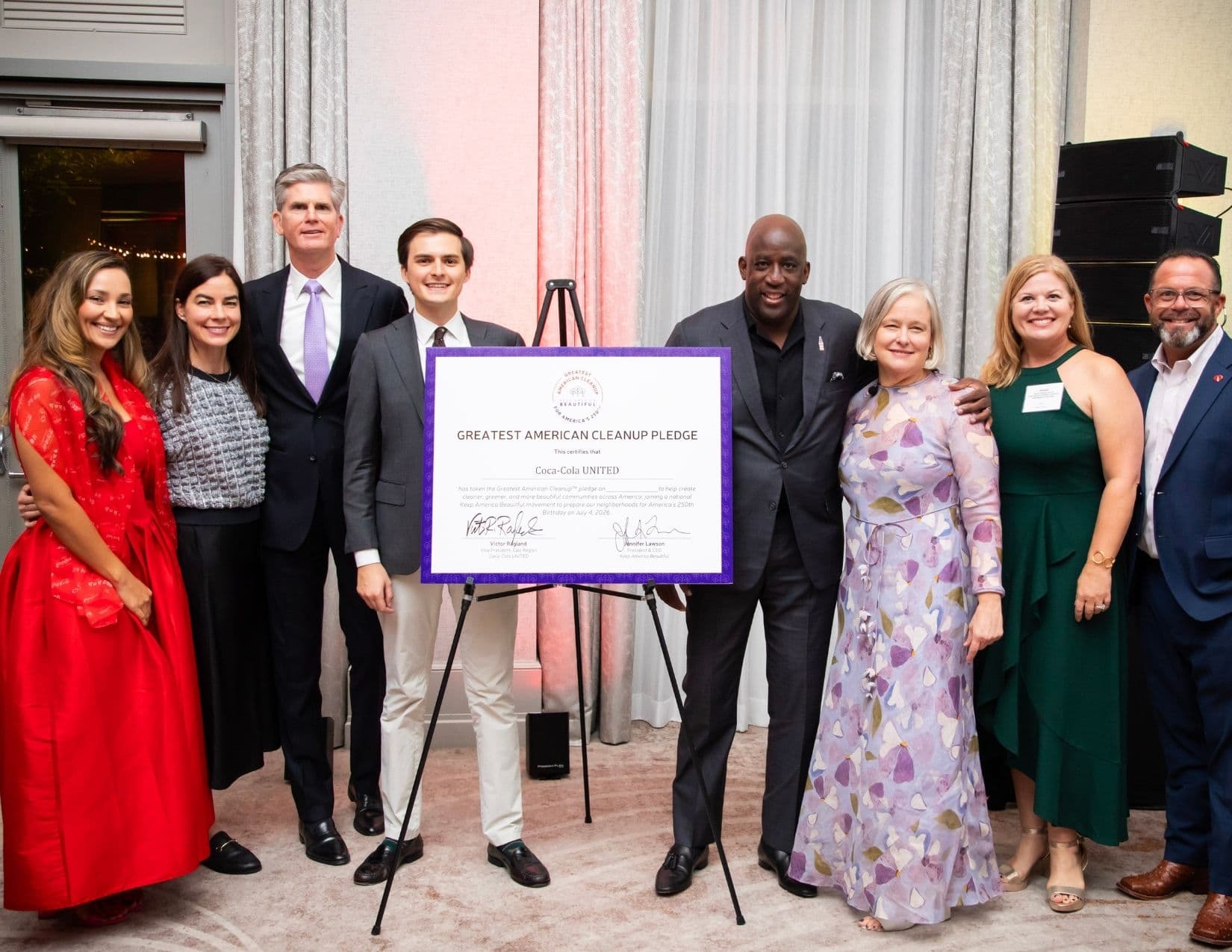 Eight people flank a large purple-bordered certificate on an easel headed GREATEST AMERICAN CLEANUP PLEDGE, certifying Coca-Cola UNITED, with two signatures at the foot; a stack of speakers stands at the right.