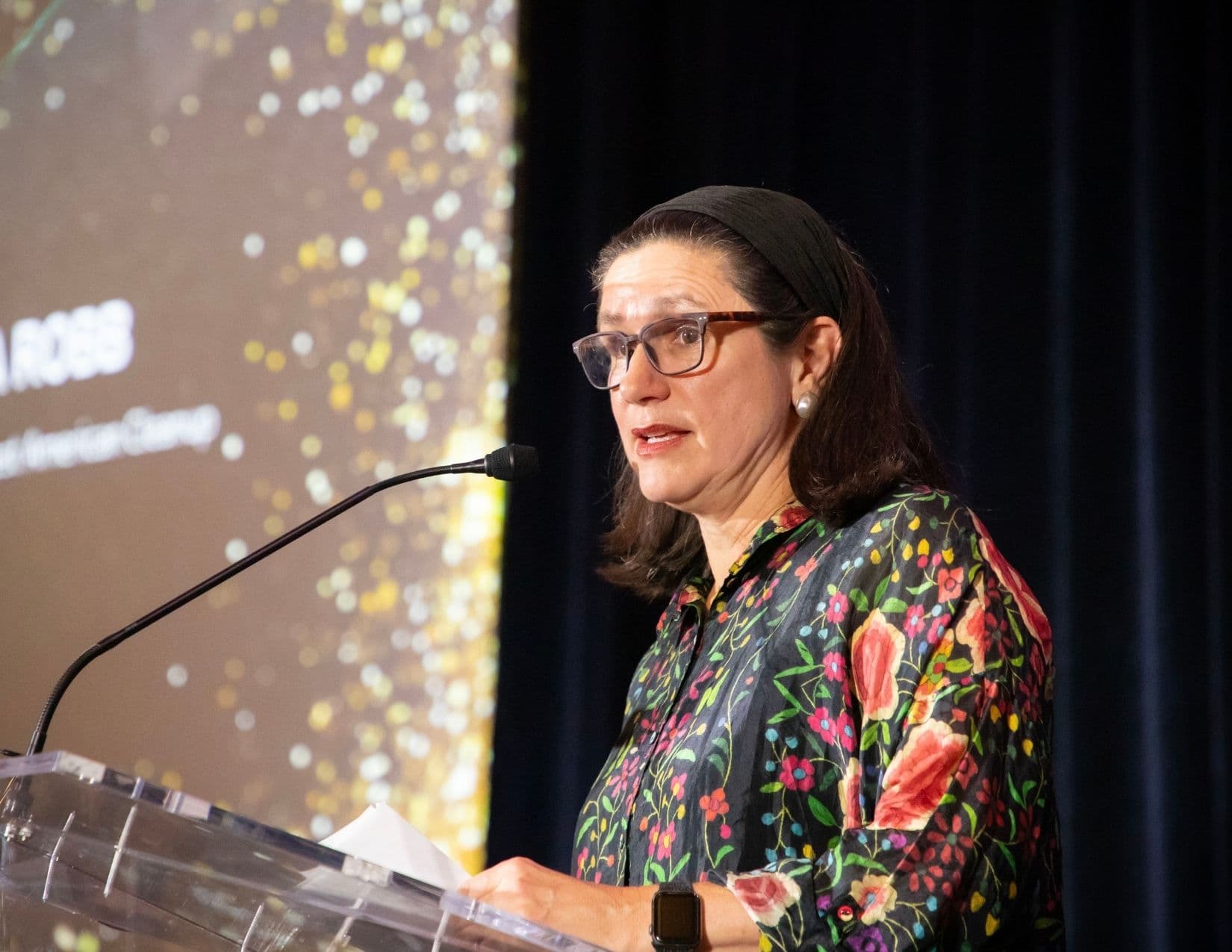 A woman in glasses, a black headband and a black blouse printed with bright folk-art flowers speaks from a clear acrylic lectern, notes in front of her and a gold-flecked projection screen behind.