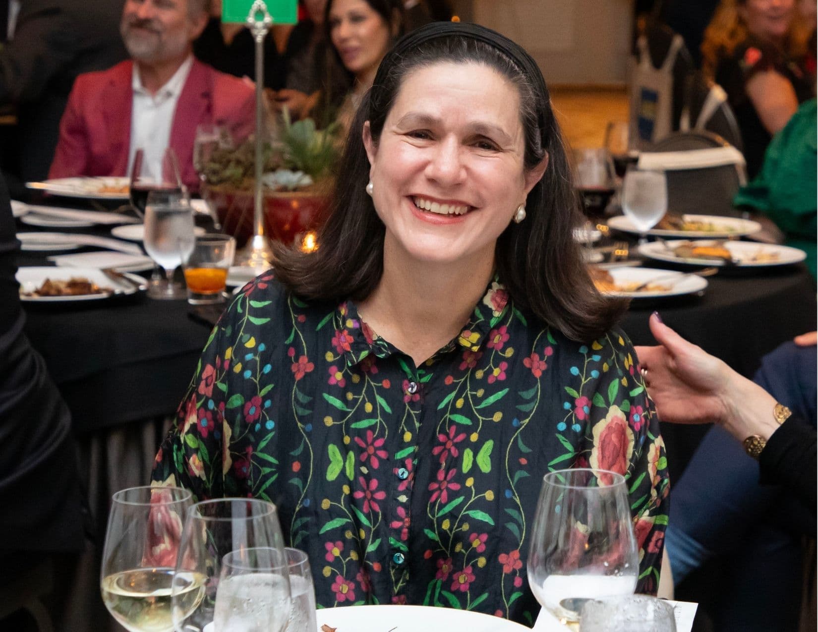 The same black floral folk-print blouse, worn by a woman laughing at a banquet table; wine glasses and a succulent centrepiece are in the foreground and a neighbour's hand rests on her shoulder.