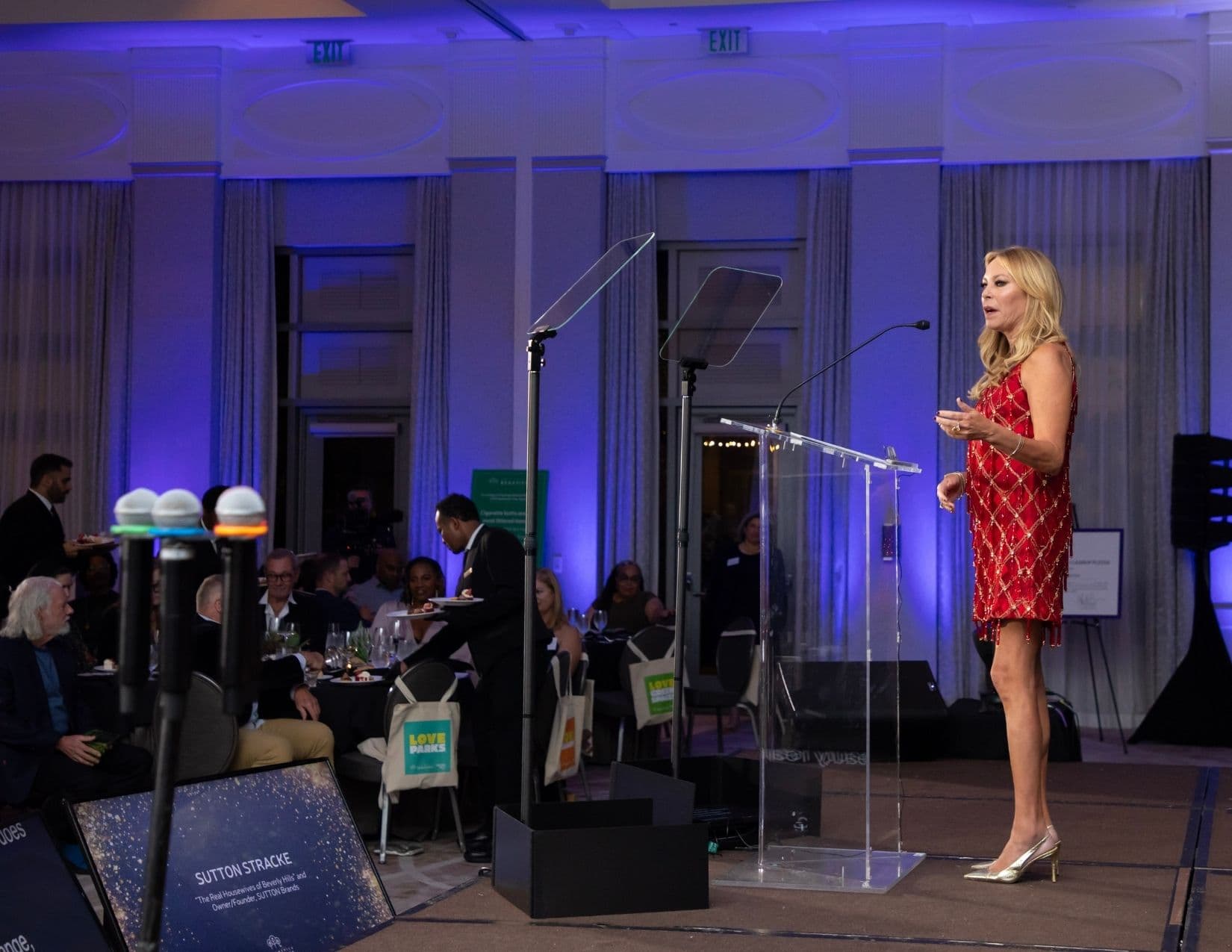 Wide shot of a blue-lit ballroom: a blonde woman in a short red and gold beaded dress speaks at a clear lectern with teleprompter screens in front of her; a floor sign at the left reads SUTTON STRACKE.