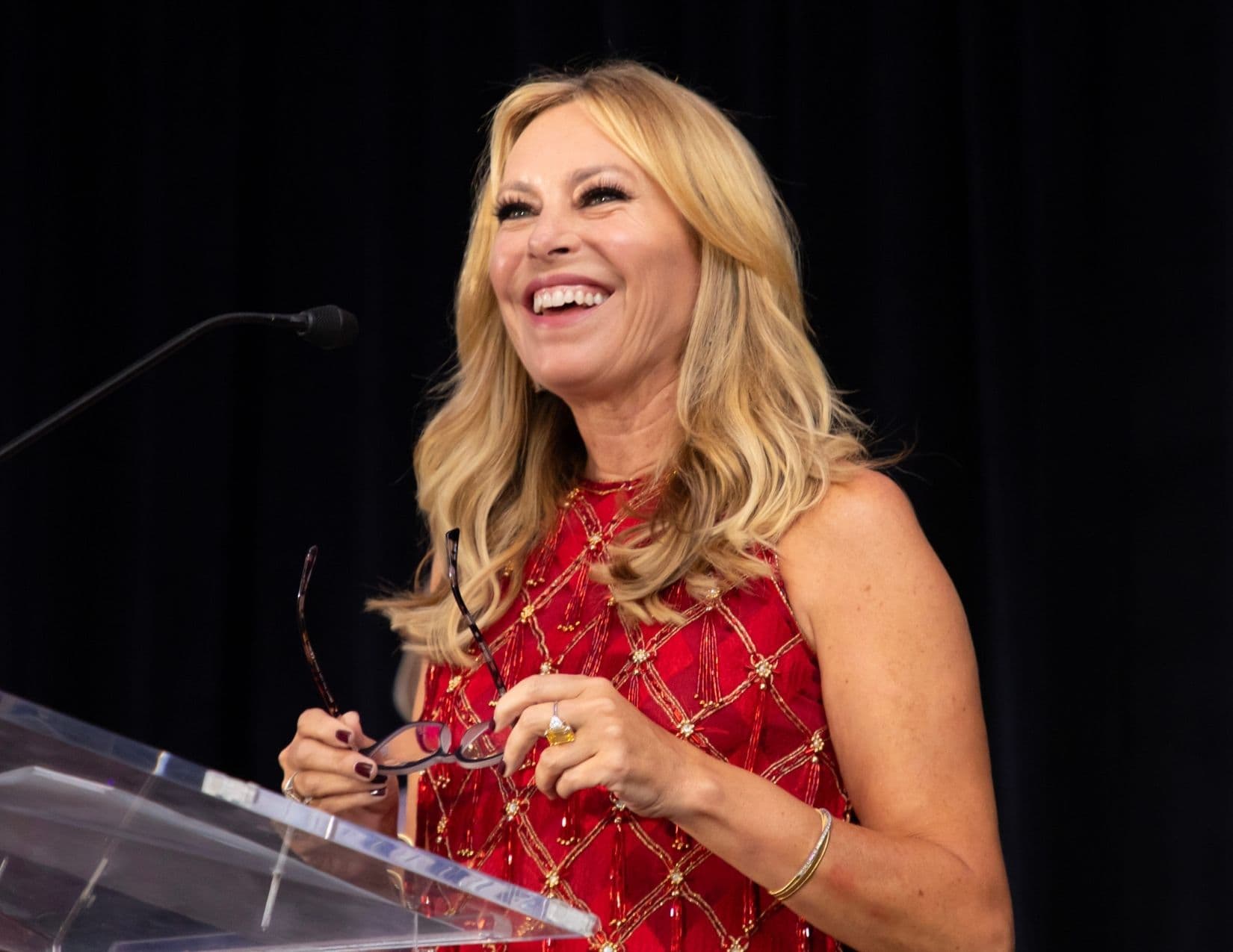 Close portrait of a laughing blonde woman in a sleeveless red dress embroidered with gold lattice work, holding folded glasses as she leans on a clear acrylic lectern against a black curtain.