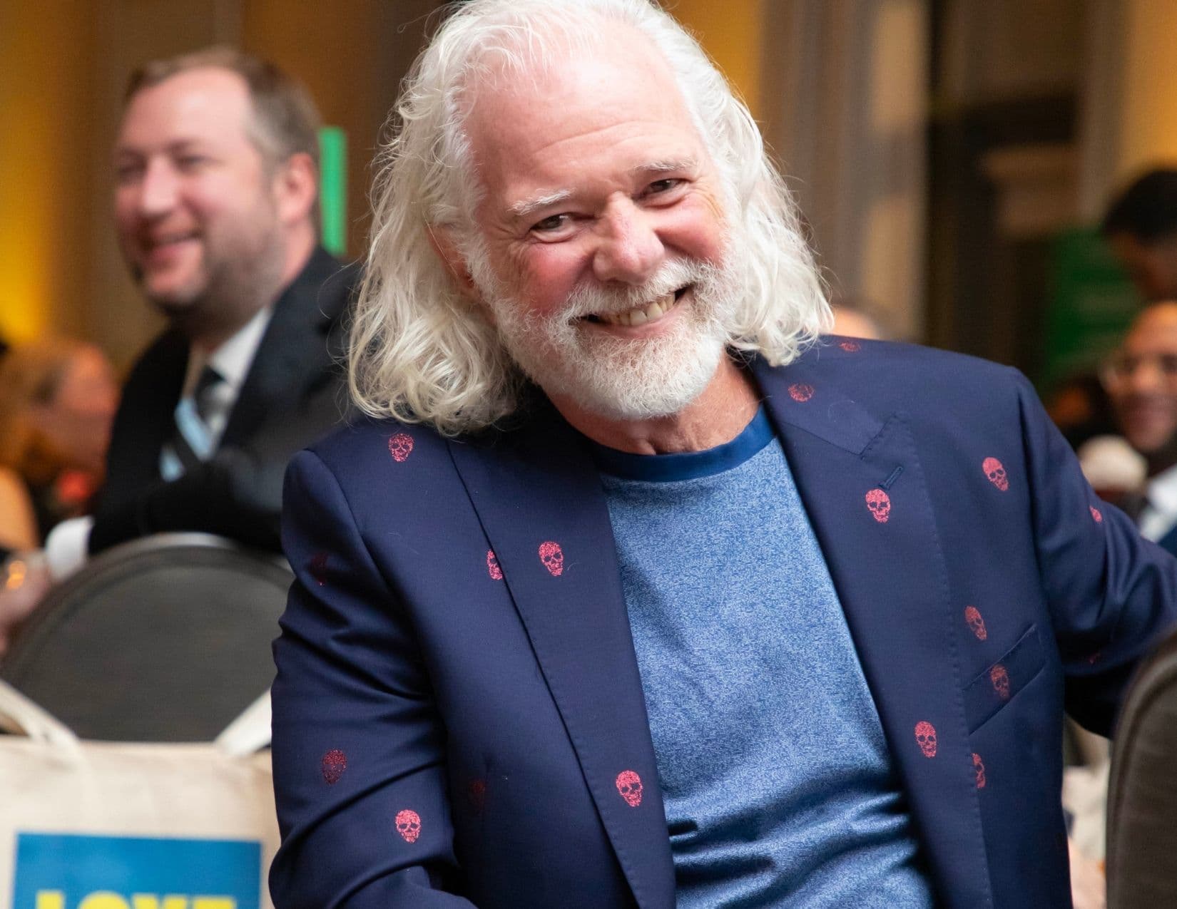 A seated man with long white hair and a full white beard smiles over his shoulder, wearing a navy blazer patterned with small red skulls over a blue crew-neck top; a canvas tote is on the chair beside him.