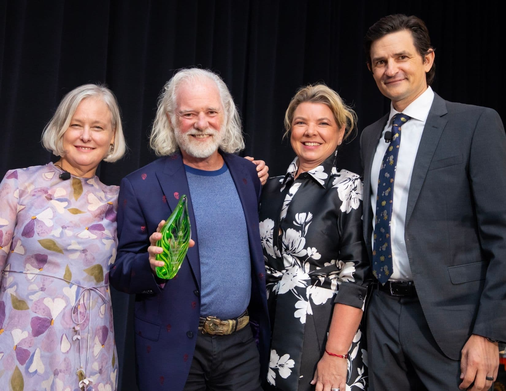 Four people on a stage in front of a black curtain; the white-bearded man in the skull-print navy blazer holds a green glass leaf-shaped award, flanked by two women and a man in a grey suit.