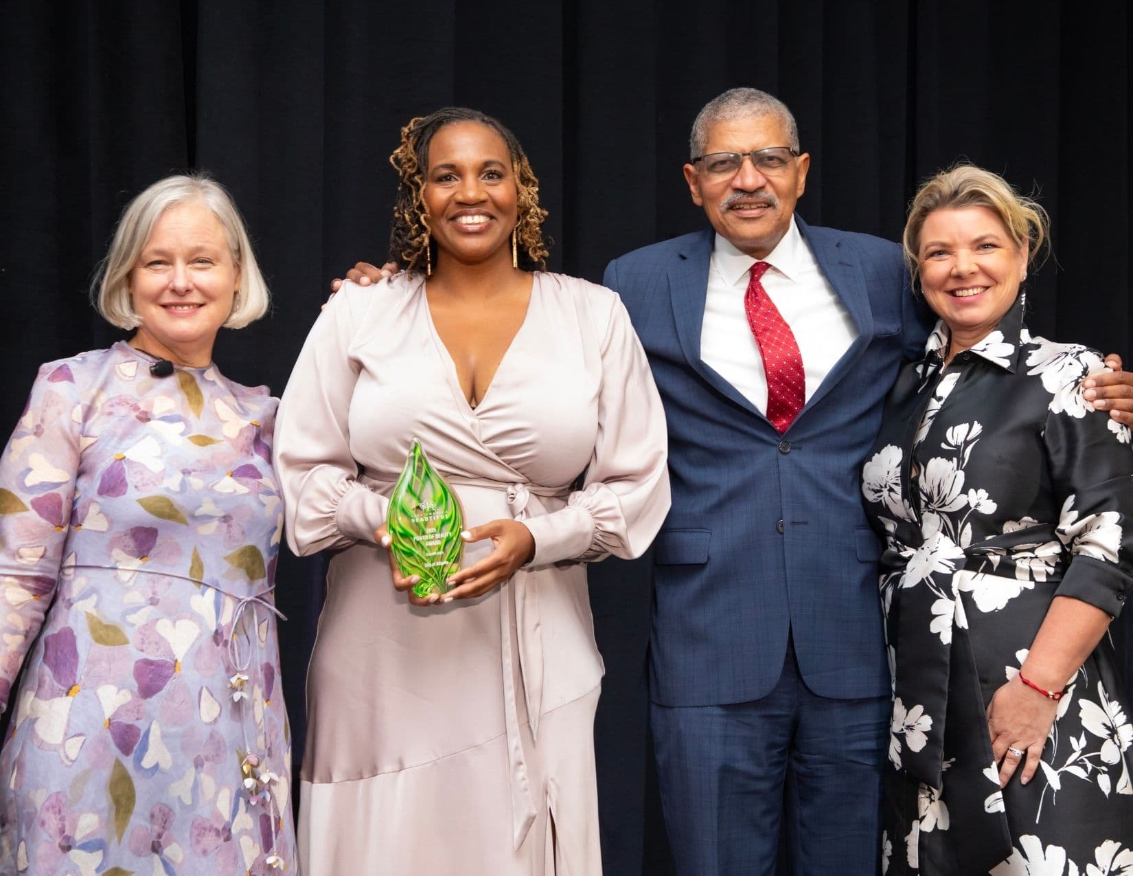Four people on stage: a woman in a blush satin wrap dress holds a green glass award reading 2025 POWER OF BEAUTY AWARD, City of Atlanta, beside a man in a navy suit with a red tie and two women in floral dresses.