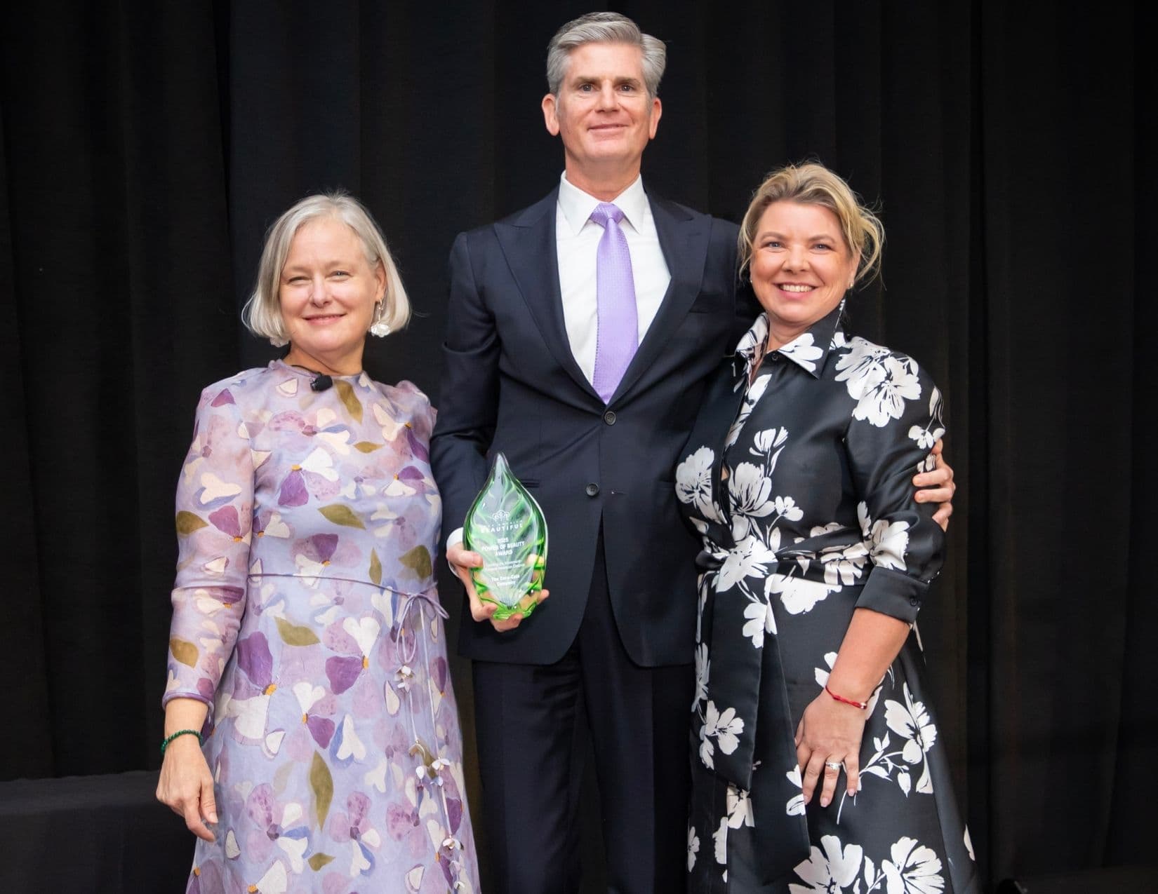 A tall man in a navy suit and lilac tie holds a green glass leaf award inscribed 2025 POWER OF BEAUTY AWARD, The Coca-Cola Company, standing between a woman in a lilac floral dress and a woman in a black and white floral dress.