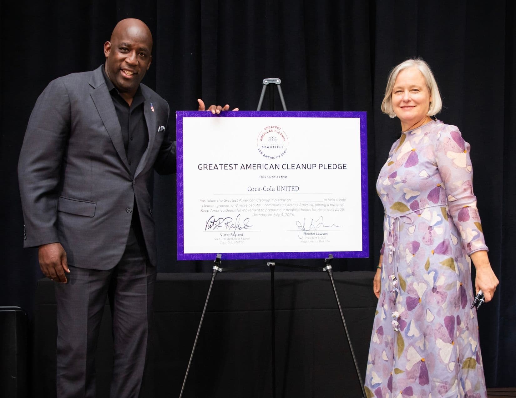 A man in a grey suit and black shirt rests a hand on a purple-framed certificate on an easel while a woman in a lilac petal-print dress stands at the other side; the board reads GREATEST AMERICAN CLEANUP PLEDGE for Coca-Cola UNITED.