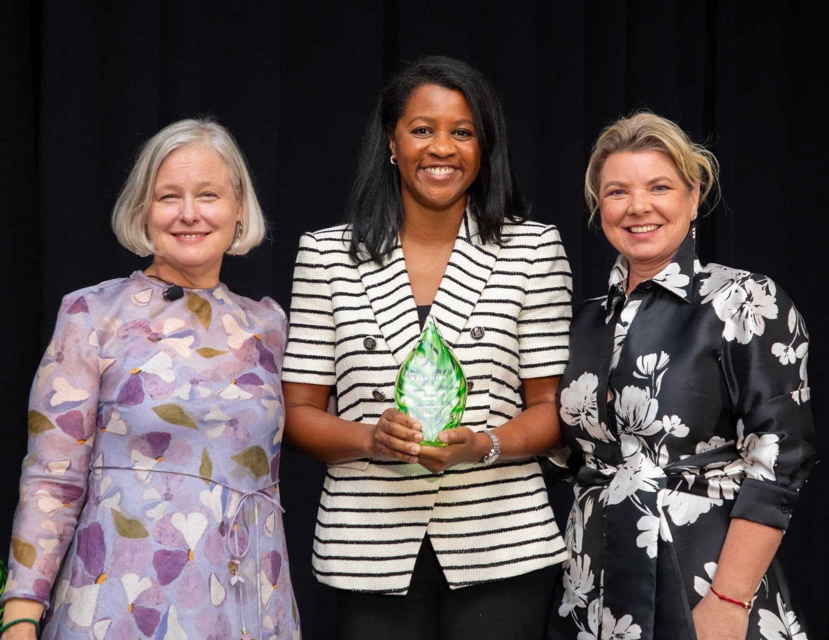 Three women stand on a stage in front of a black curtain; the one in the centre, in a cream blazer with fine black stripes, holds a green glass leaf-shaped Power of Beauty award in both hands.
