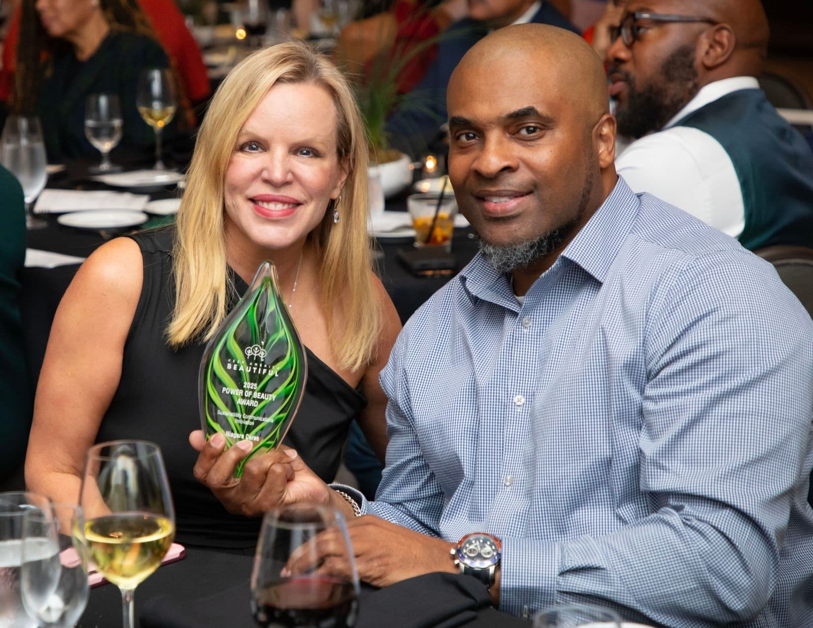 Seated at a banquet table, a blonde woman in a one-shoulder black dress holds up a green glass award reading 2025 POWER OF BEAUTY AWARD, Sustainability Communications Innovation, Niagara Cares, next to a man in a blue checked shirt.