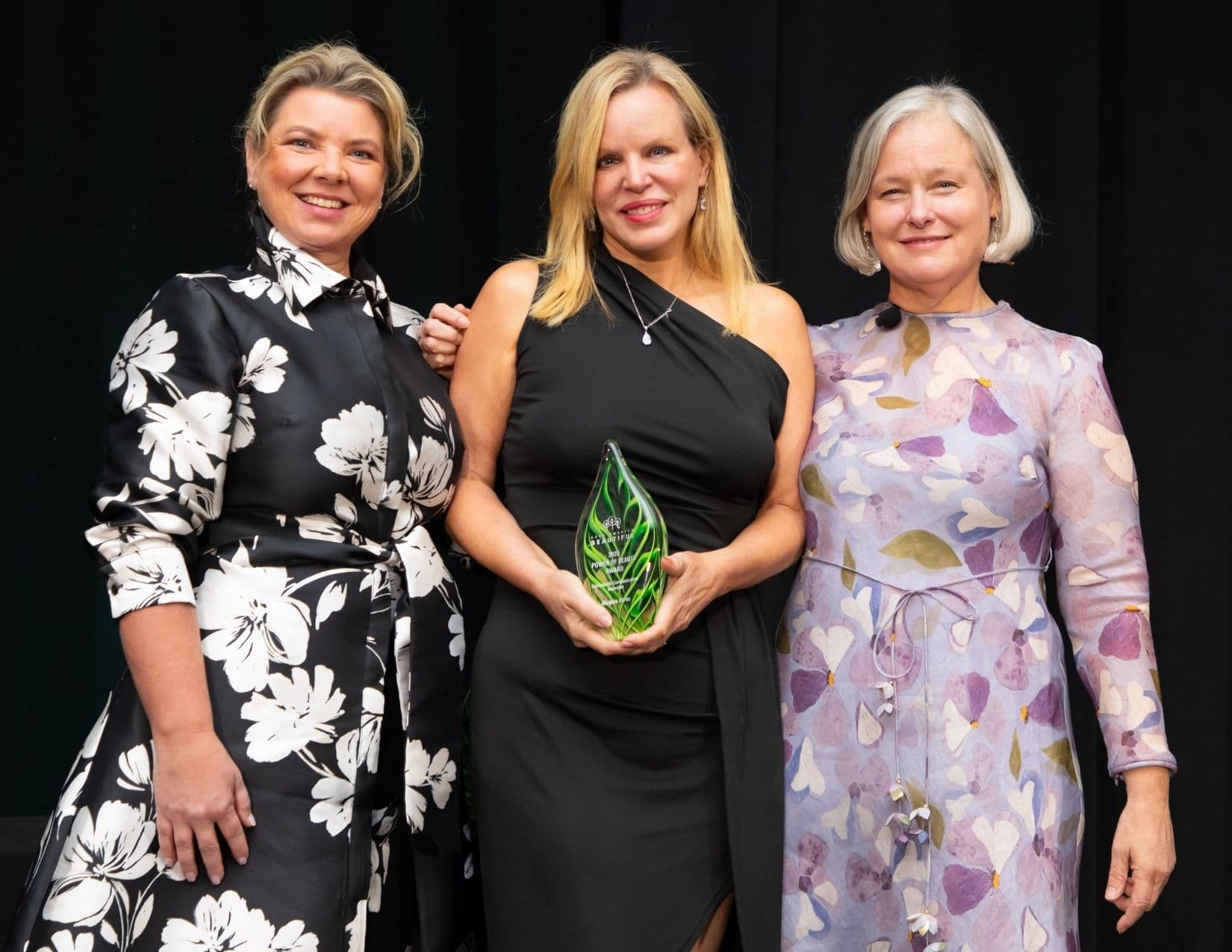 Three women stand on stage; the blonde in the centre, in a one-shoulder black gown, cradles the green glass Niagara Cares award, with a woman in a black and white floral dress at left and one in a lilac petal print at right.