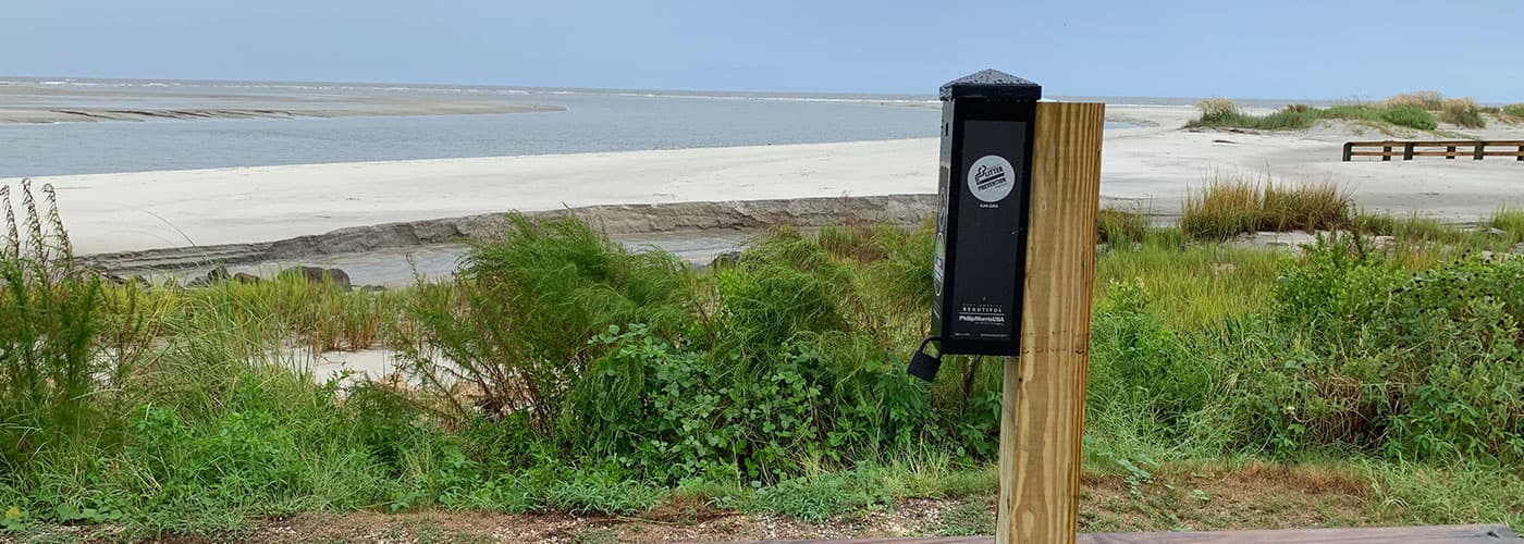 A black cigarette-butt receptacle mounted on a wooden post at the edge of a beach boardwalk, its label carrying a Keep America Beautiful mark and Philip Morris USA text, with dune grass, pale sand and calm water behind.
