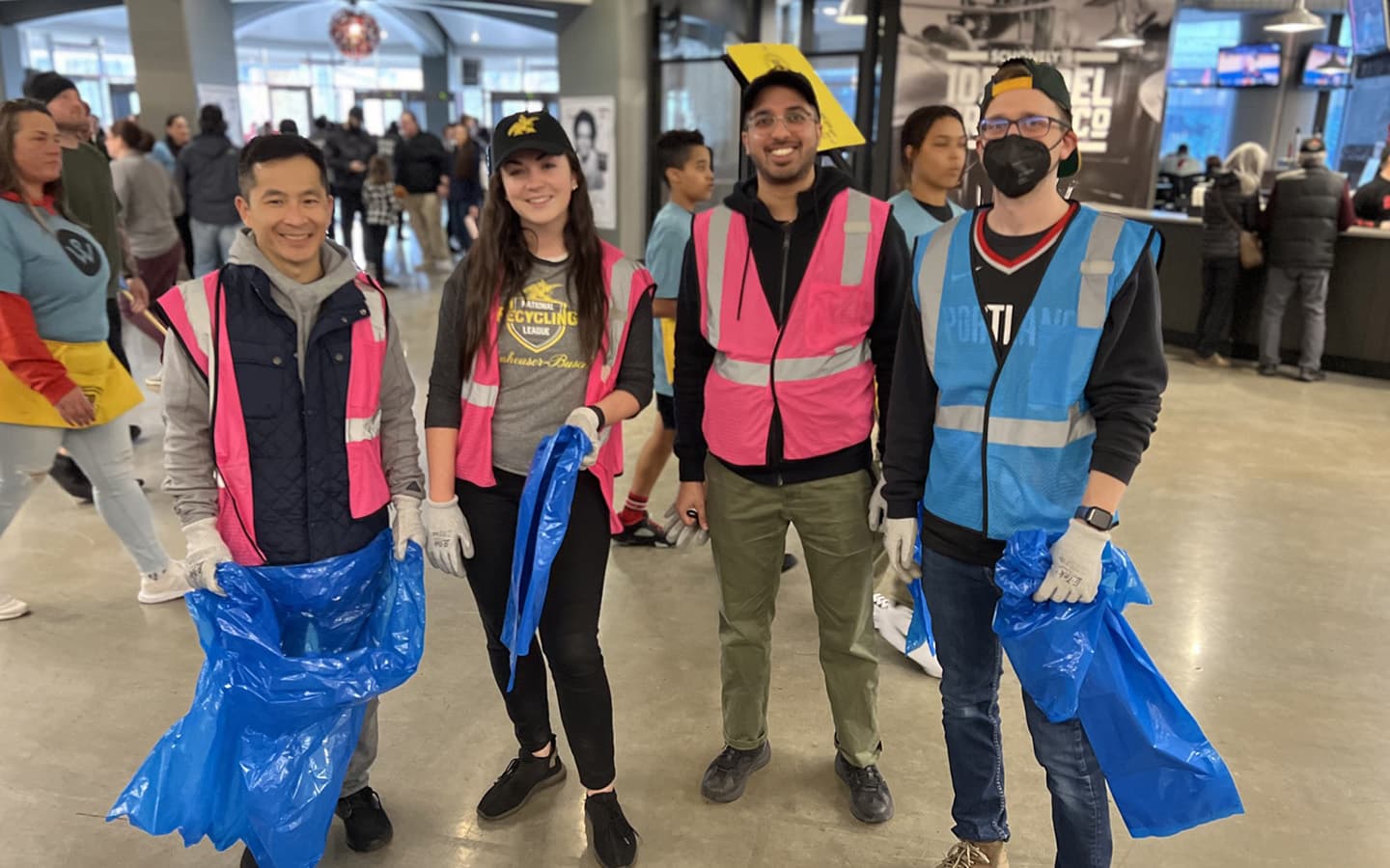 Four volunteers in pink and blue hi-vis vests and work gloves stand in a stadium concourse holding open blue rubbish sacks; one wears a National Recycling League t-shirt and another a black face mask and backwards cap.