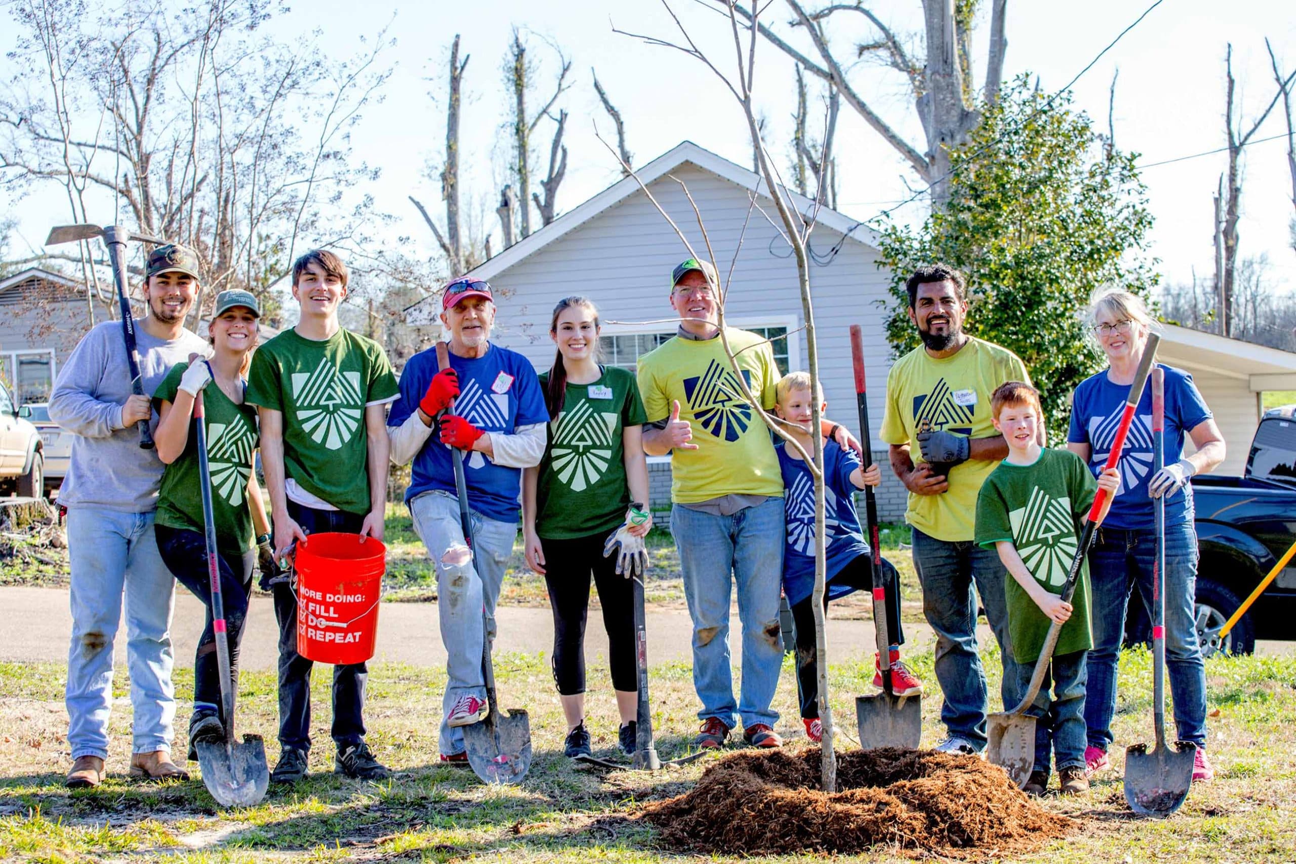 Eleven volunteers of mixed ages, in green, yellow and blue t-shirts printed with a stylised tree emblem, pose with shovels and a red bucket around a freshly mulched sapling in a front garden; a red bucket reads MORE DOING: FILL, DO, REPEAT.
