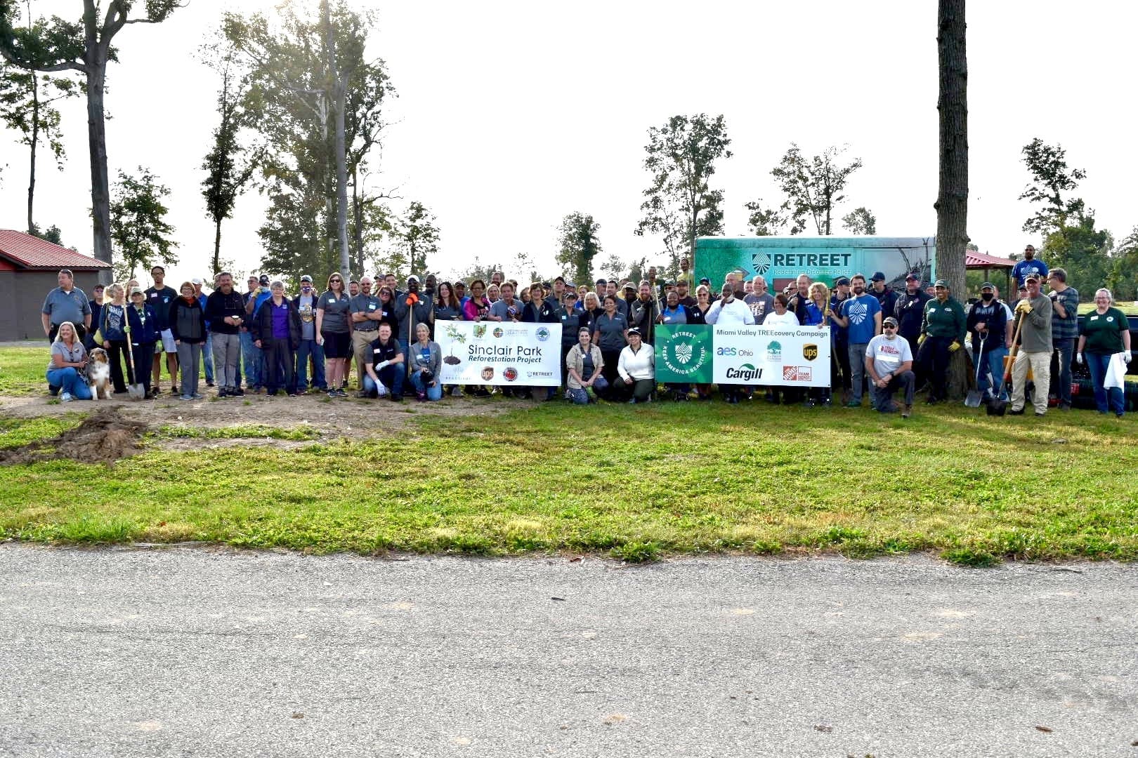 A very large crowd of volunteers lines up on grass in front of a green box trailer lettered RETREET, holding two banners: Sinclair Park Reforestation Project, and Miami Valley TREEcovery Campaign with AES Ohio, UPS, Cargill and The Home Depot logos.